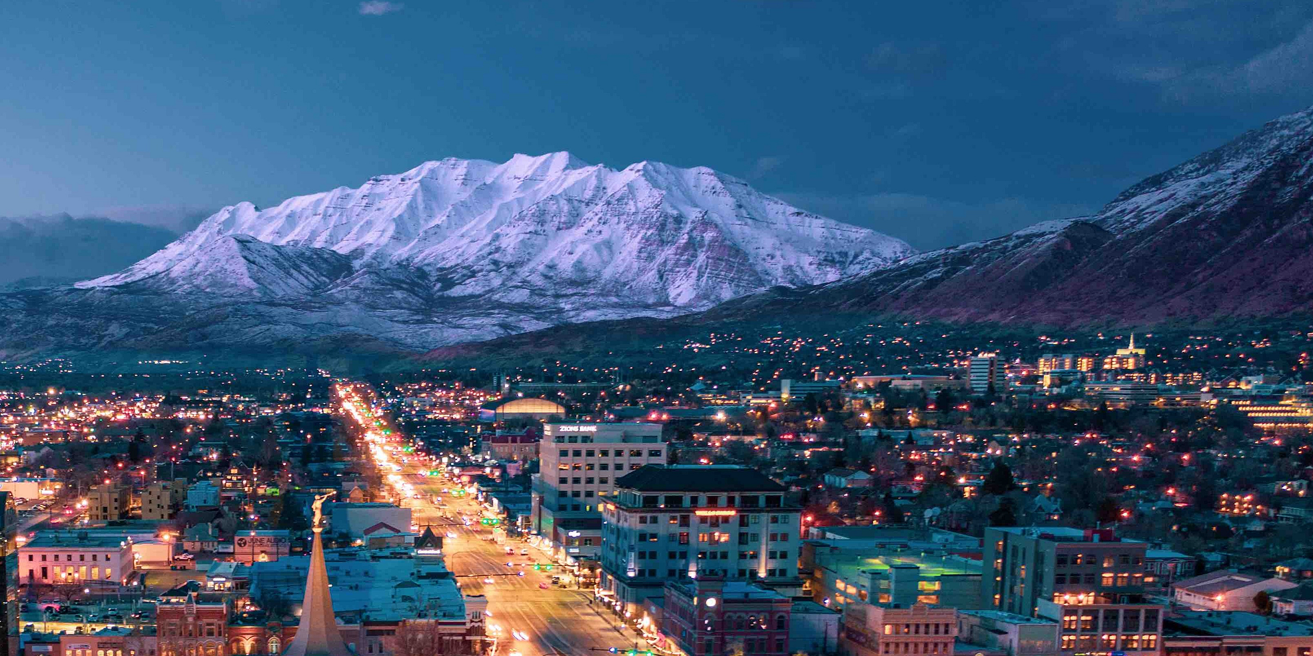 Breathtaking twilight cityscape with vibrant urban lights against a backdrop of majestic snow-covered mountains.