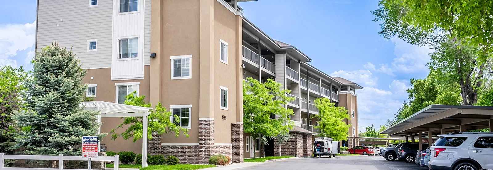 Charming apartment complex with lush greenery, ample parking, and scenic mountain views under a bright blue sky.