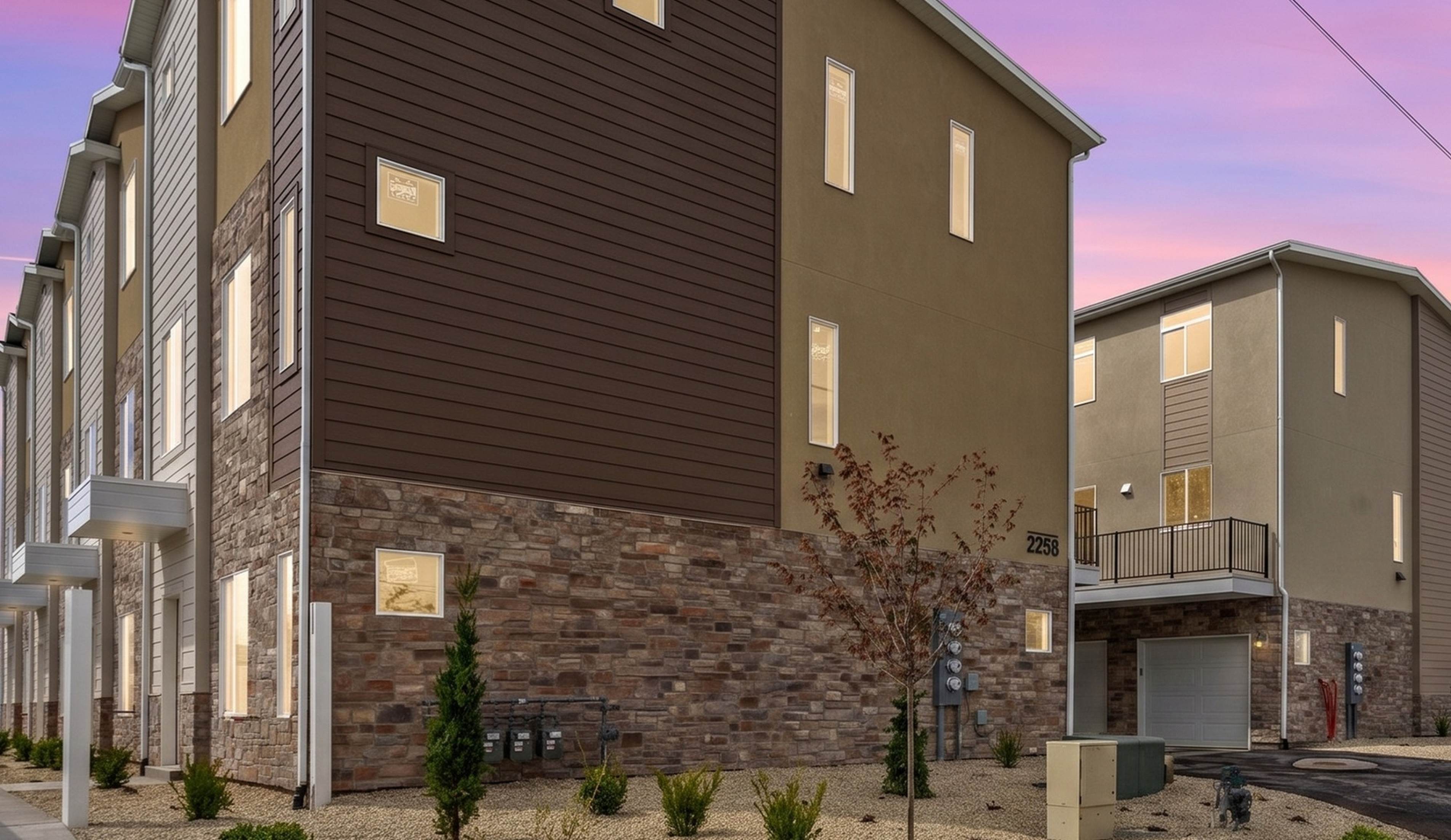 Modern townhome exterior with stylish stone accents and landscaped gardens against a stunning sunset backdrop.