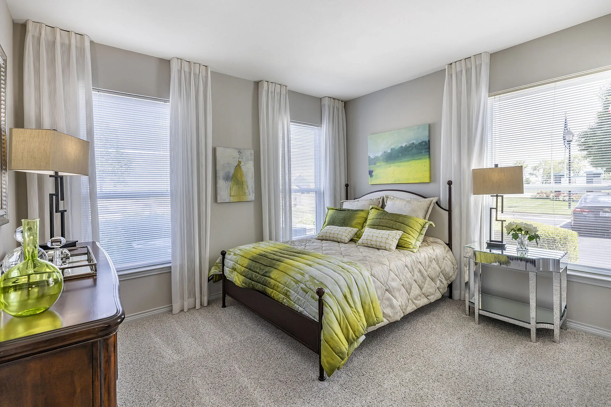 Bright and elegant bedroom featuring a plush bed with green accents, modern decor, and large windows inviting natural light.