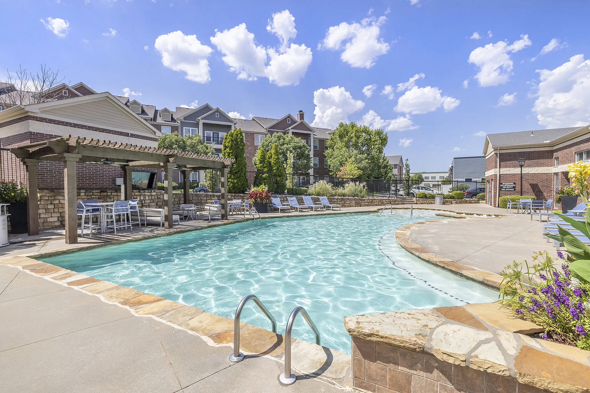 Inviting outdoor pool area with stylish lounge chairs and a shaded pergola, surrounded by beautiful brick buildings and lush greenery under a bright blue sky.