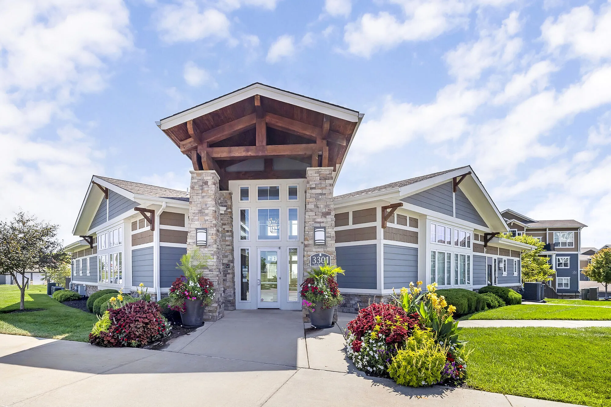 Elegant entrance of a modern apartment complex featuring stone columns, large windows, and vibrant landscaping under a clear blue sky.