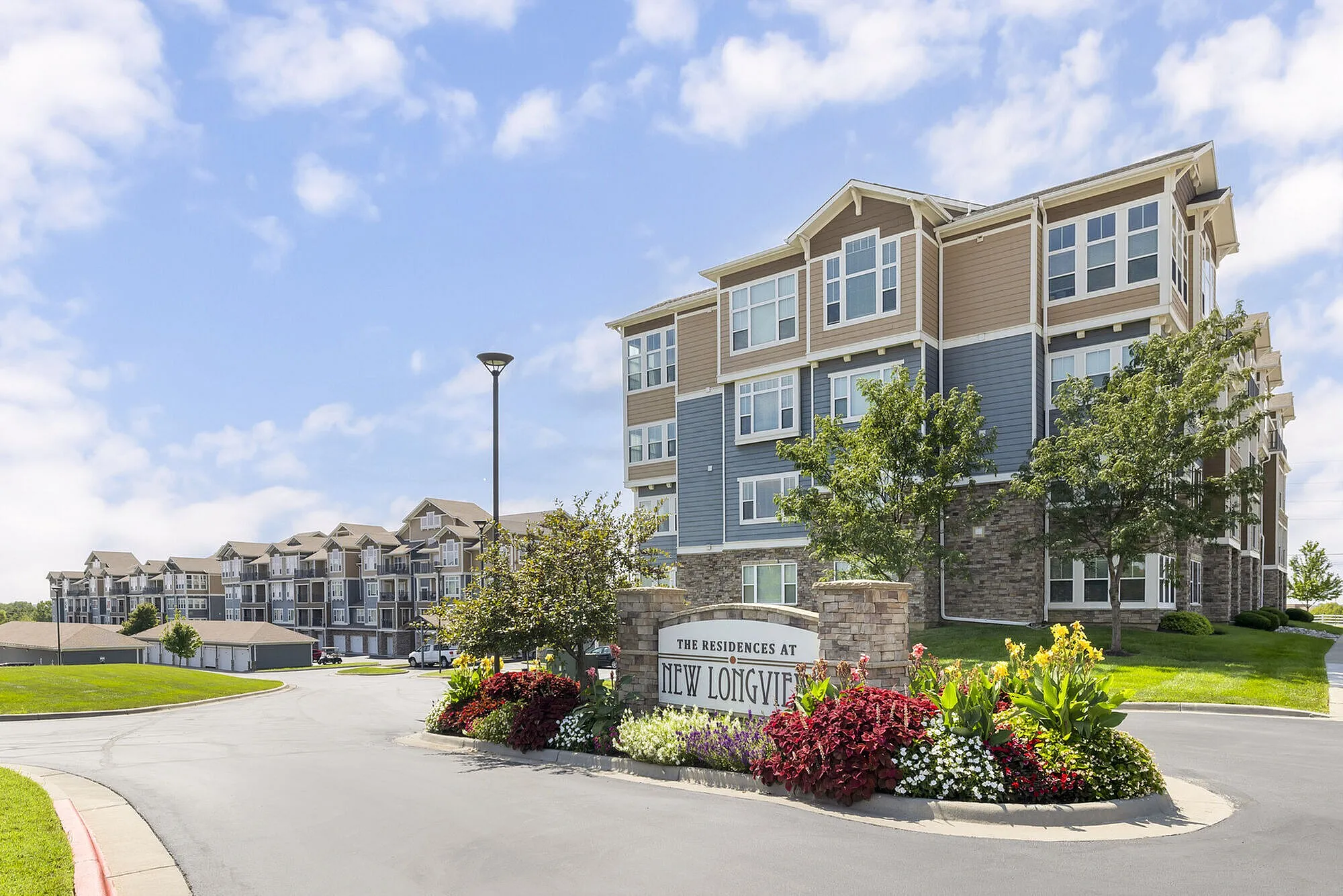Charming multi-story apartment complex with modern architecture, surrounded by lush landscaping and vibrant flower beds under a clear blue sky.