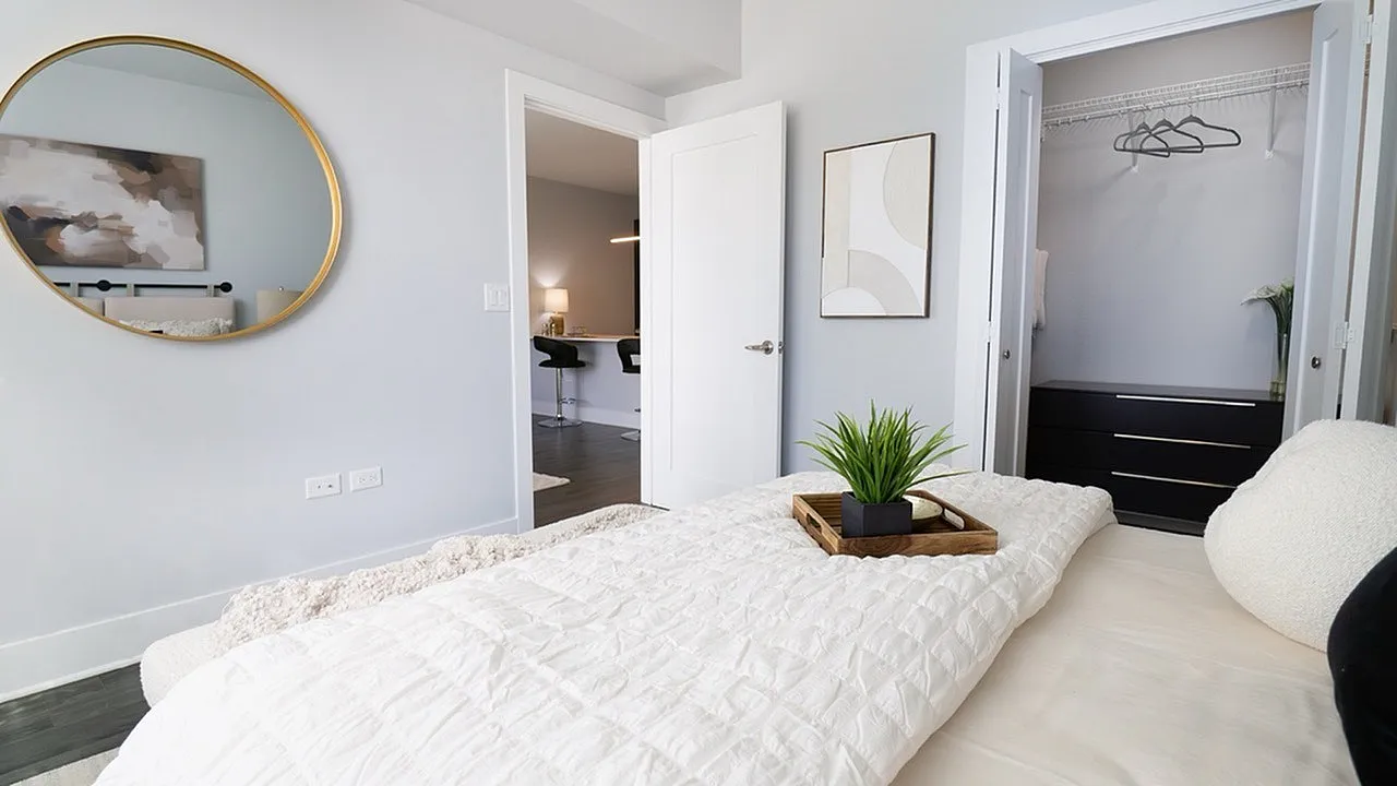 Modern bedroom with a cozy aesthetic, featuring a stylish round mirror, sleek closet space, and inviting decor with a glimpse into a well-lit study area.