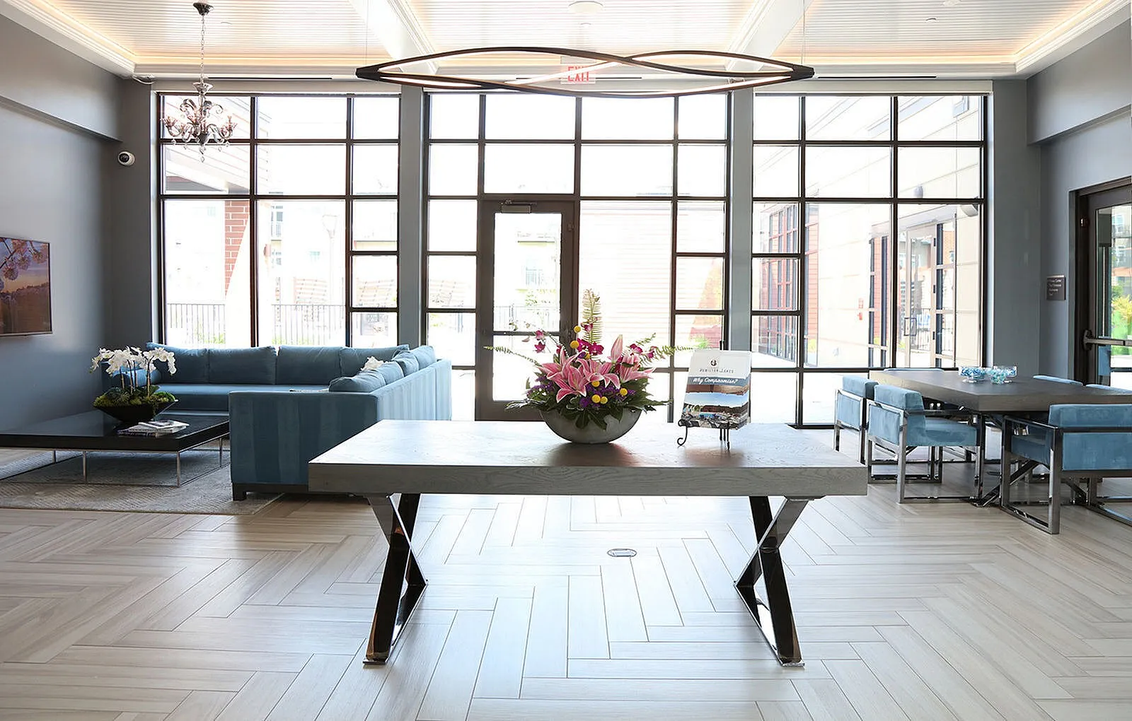 Bright modern living space with large windows, elegant chandelier, and stylish seating area featuring a plush blue sofa and sleek dining table.
