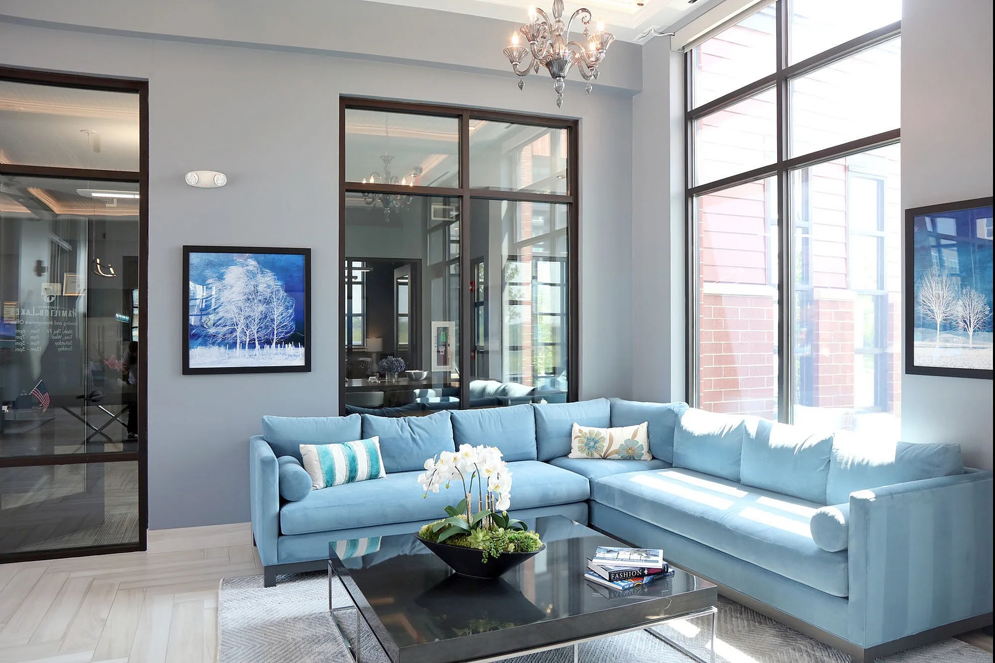Bright and elegant living room with large windows, featuring a plush blue sectional sofa, chic chandelier, and modern artwork.