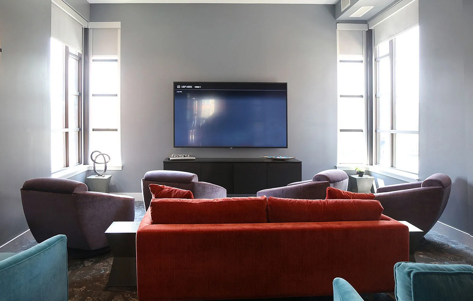 Chic living room with a modern flat-screen TV, vibrant red sofa, and cozy armchairs, flooded with natural light from large windows.
