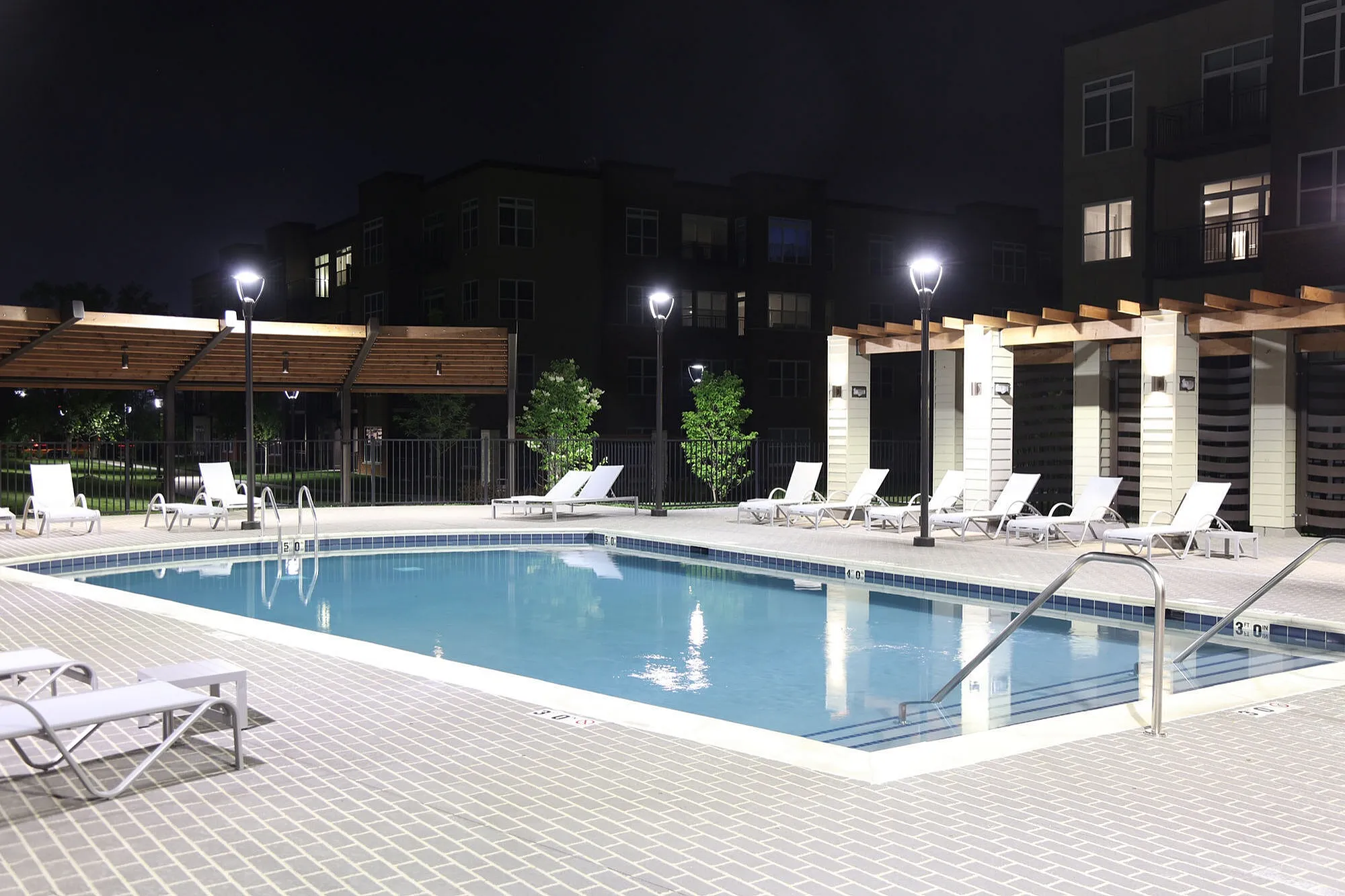 Elegant night view of a well-lit swimming pool area with modern lounge chairs and pergola accents, perfect for relaxation.