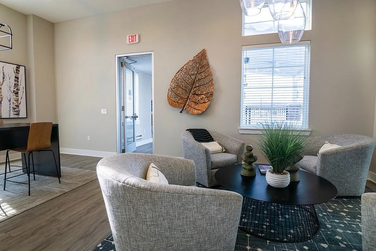 Modern lounge area with elegant grey armchairs, a chic black coffee table, and contemporary leaf artwork on the wall, filled with natural light from a large window.