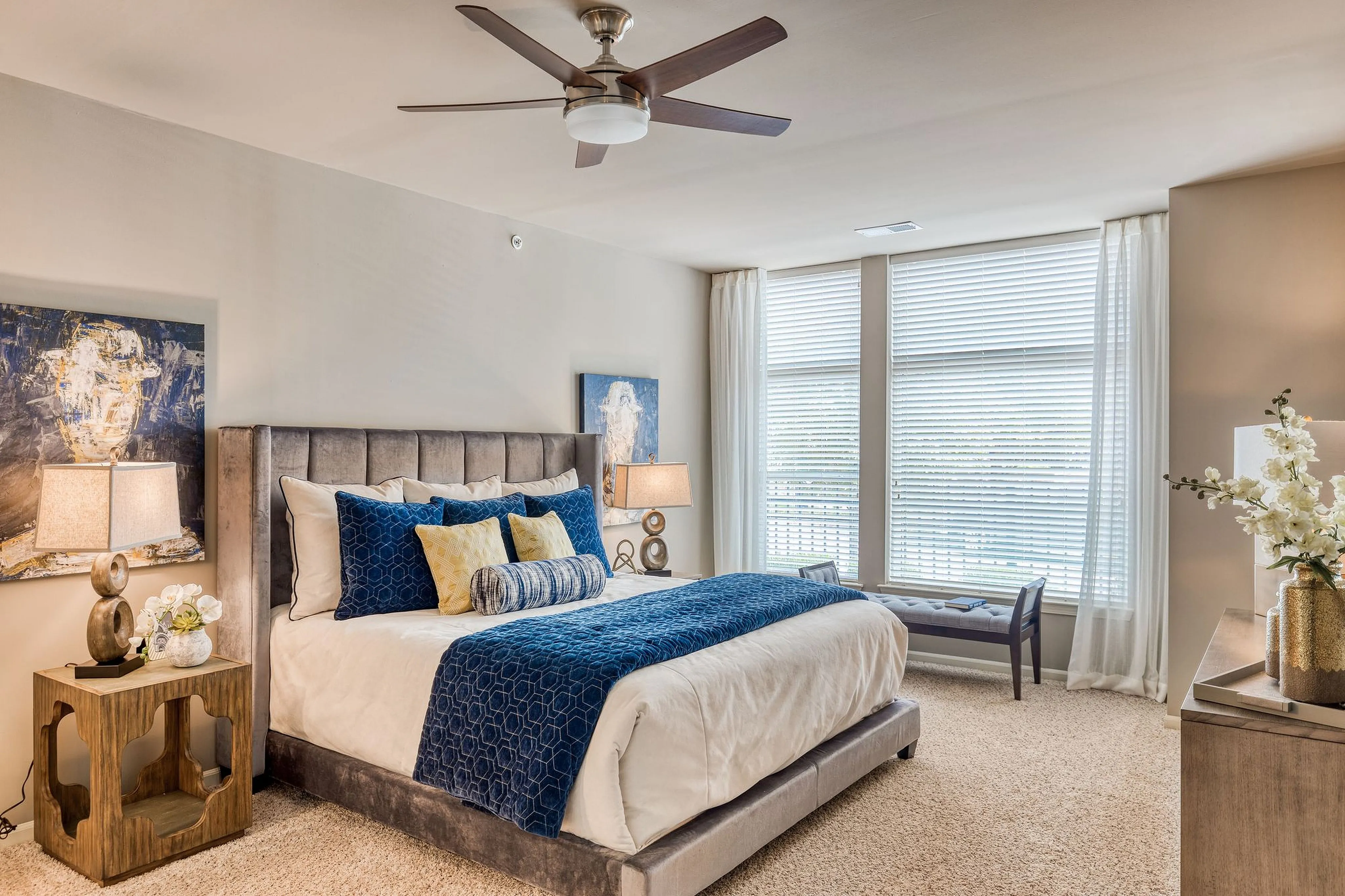 Elegant bedroom featuring a plush bed with stylish blue accents, modern decor, and large windows inviting natural light.