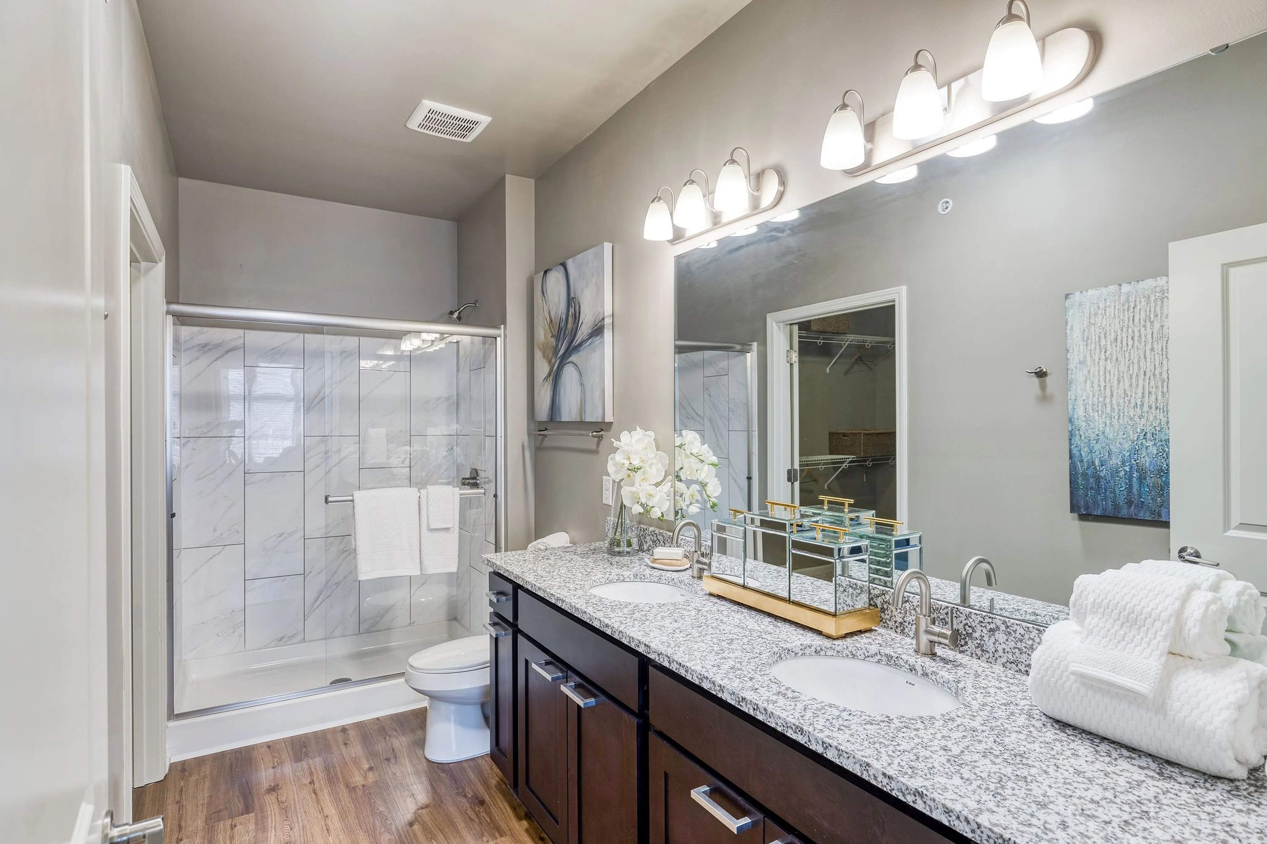Elegant bathroom with granite countertops, dual sinks, and a spacious glass-enclosed shower. Features warm wood flooring and tasteful artwork for a relaxing ambiance.