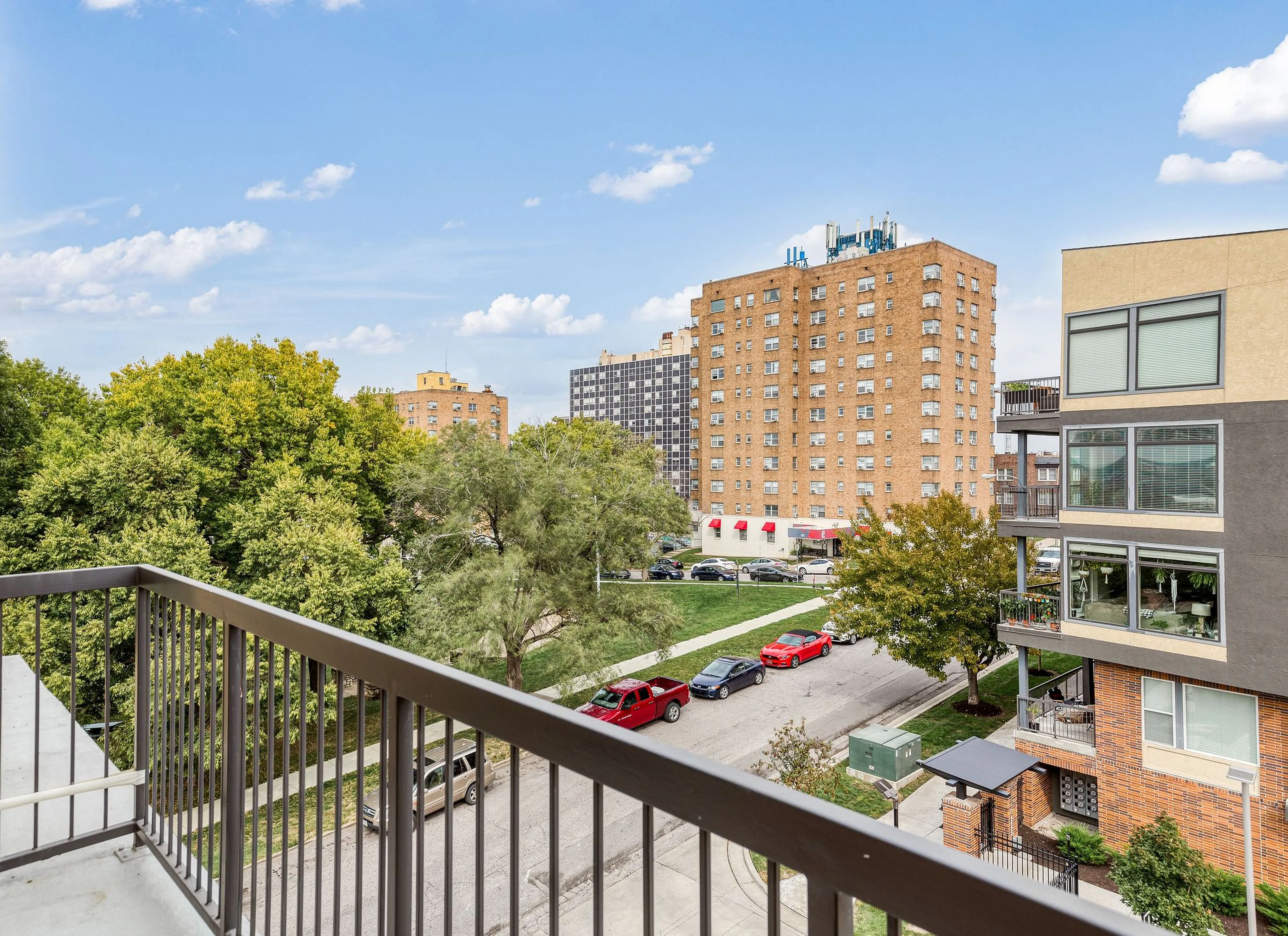 Charming cityscape view from a balcony overlooking lush trees and towering apartment buildings under a bright blue sky.