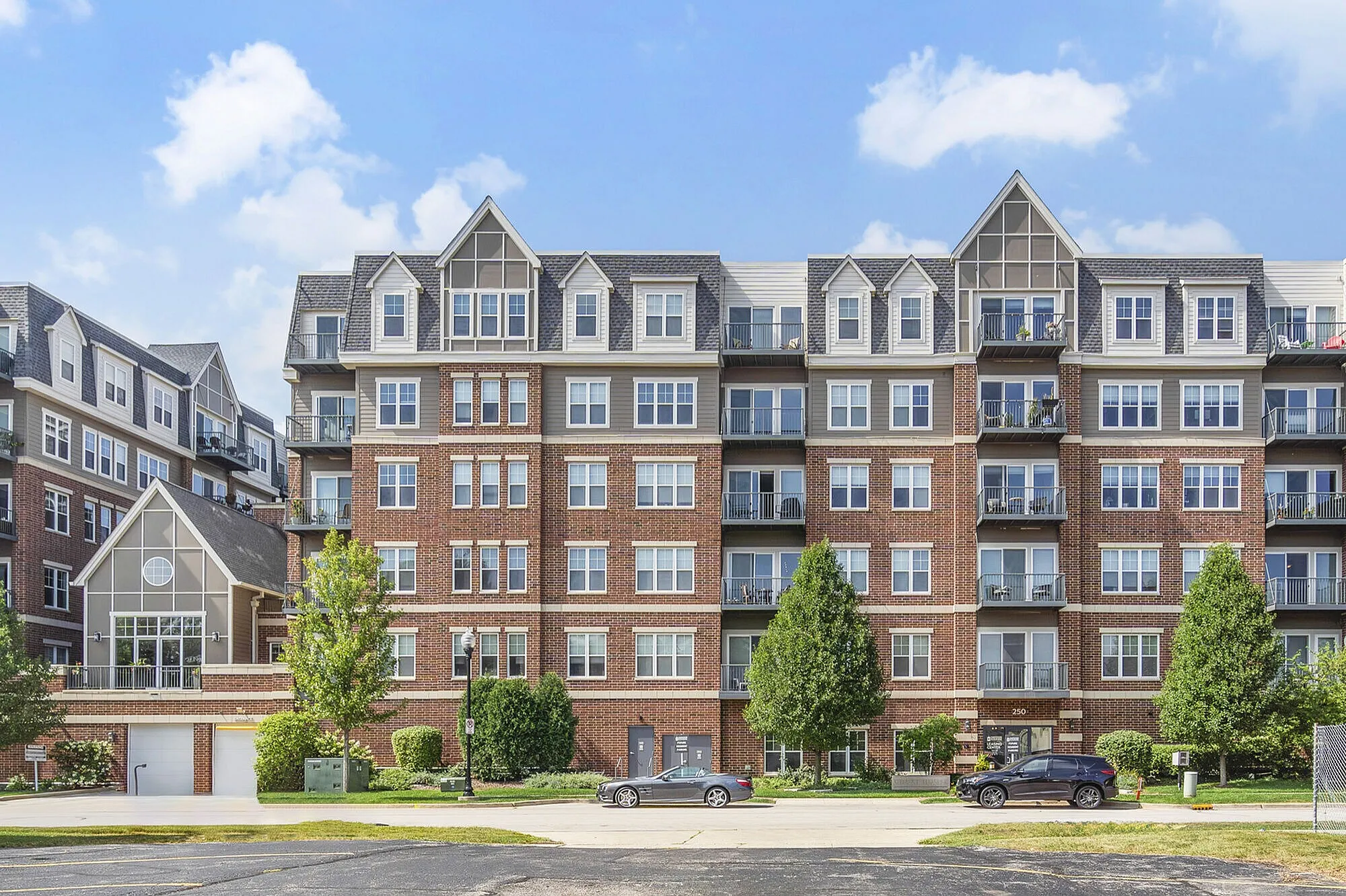 Elegant multi-story apartment building with modern brick design, large windows, and spacious balconies, surrounded by lush greenery and clear blue skies.