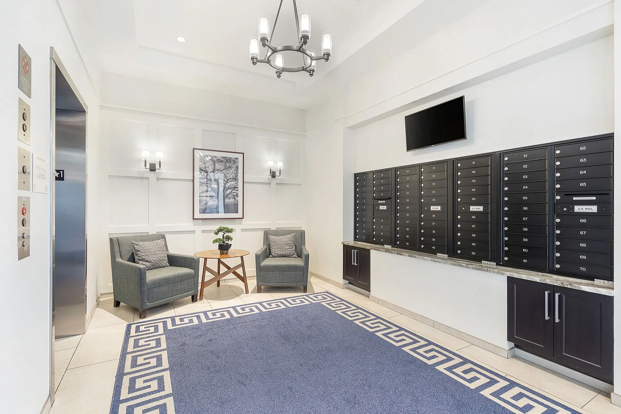 Elegant apartment lobby featuring a modern chandelier, cozy seating with two armchairs and a table, stylish wall sconces, and an organized mailroom with black mailboxes.