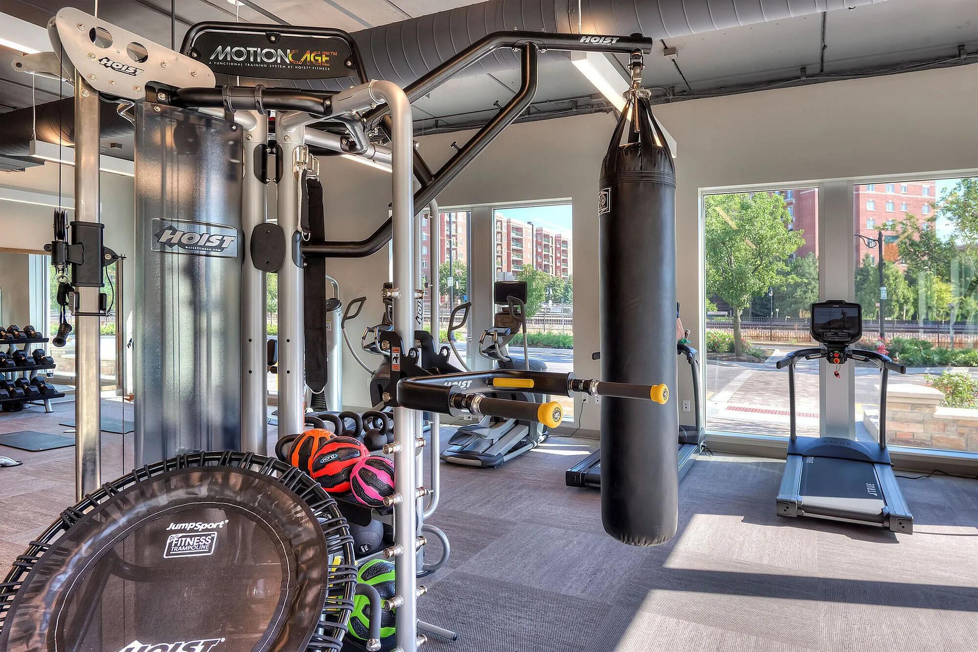 Modern fitness center with state-of-the-art equipment including a functional training system, punching bag, and treadmill, set in a bright space with large windows offering a view of the urban landscape.