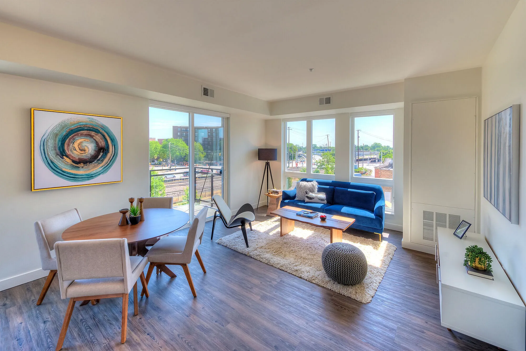 Bright and modern living space featuring a cozy blue sofa, sleek wooden dining table, and large windows with a city view.