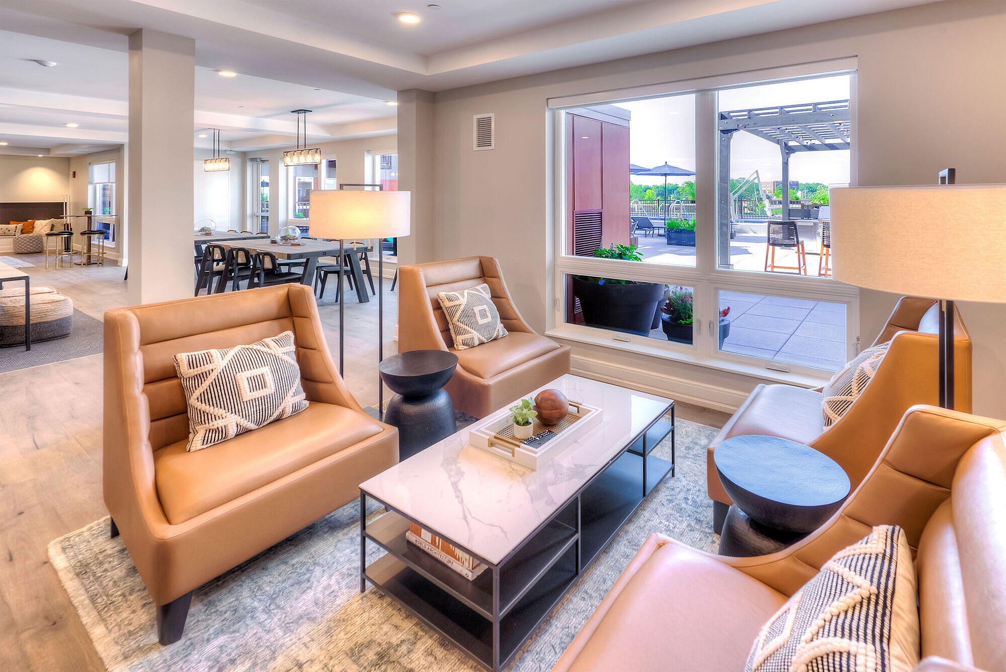 Elegant and spacious living area with stylish tan leather chairs, a sleek marble coffee table, and abundant natural light from large windows overlooking a vibrant outdoor patio.