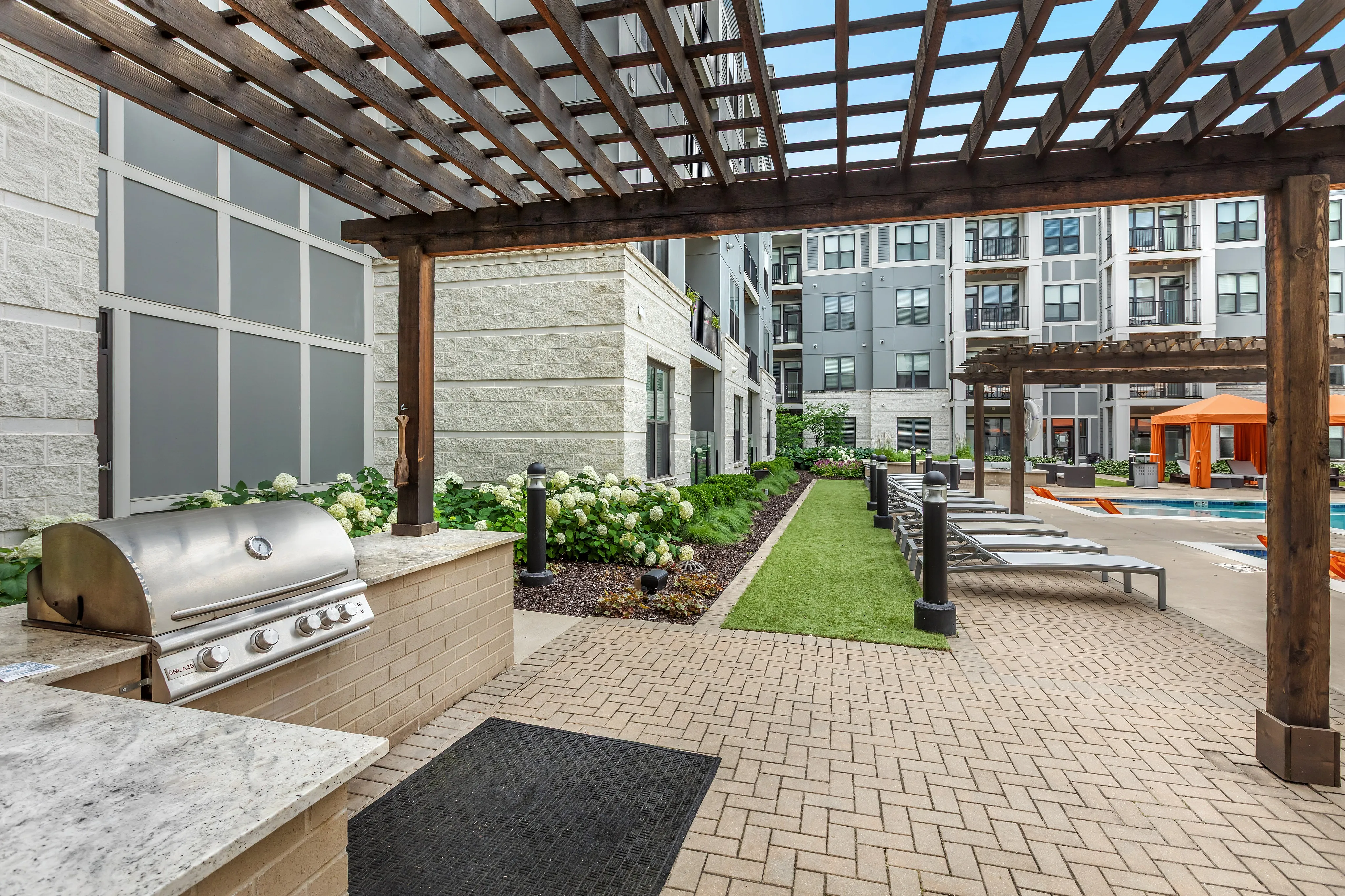 Luxurious outdoor apartment amenities featuring a stylish pergola, modern grill area, and inviting poolside loungers with vibrant orange cabanas.
