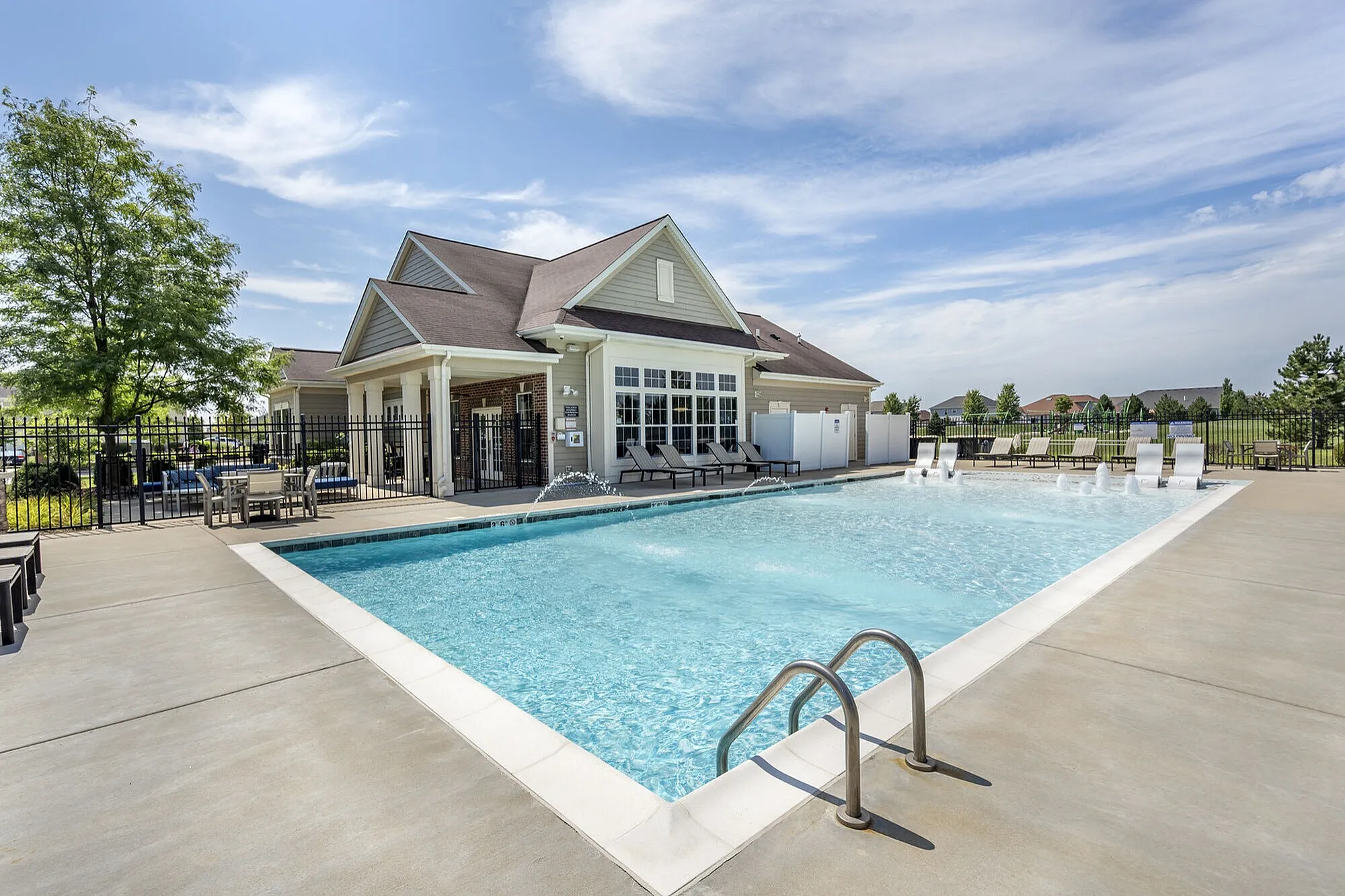 Inviting outdoor swimming pool area with modern patio furniture and pristine clubhouse, surrounded by lush landscaping under a blue sky.