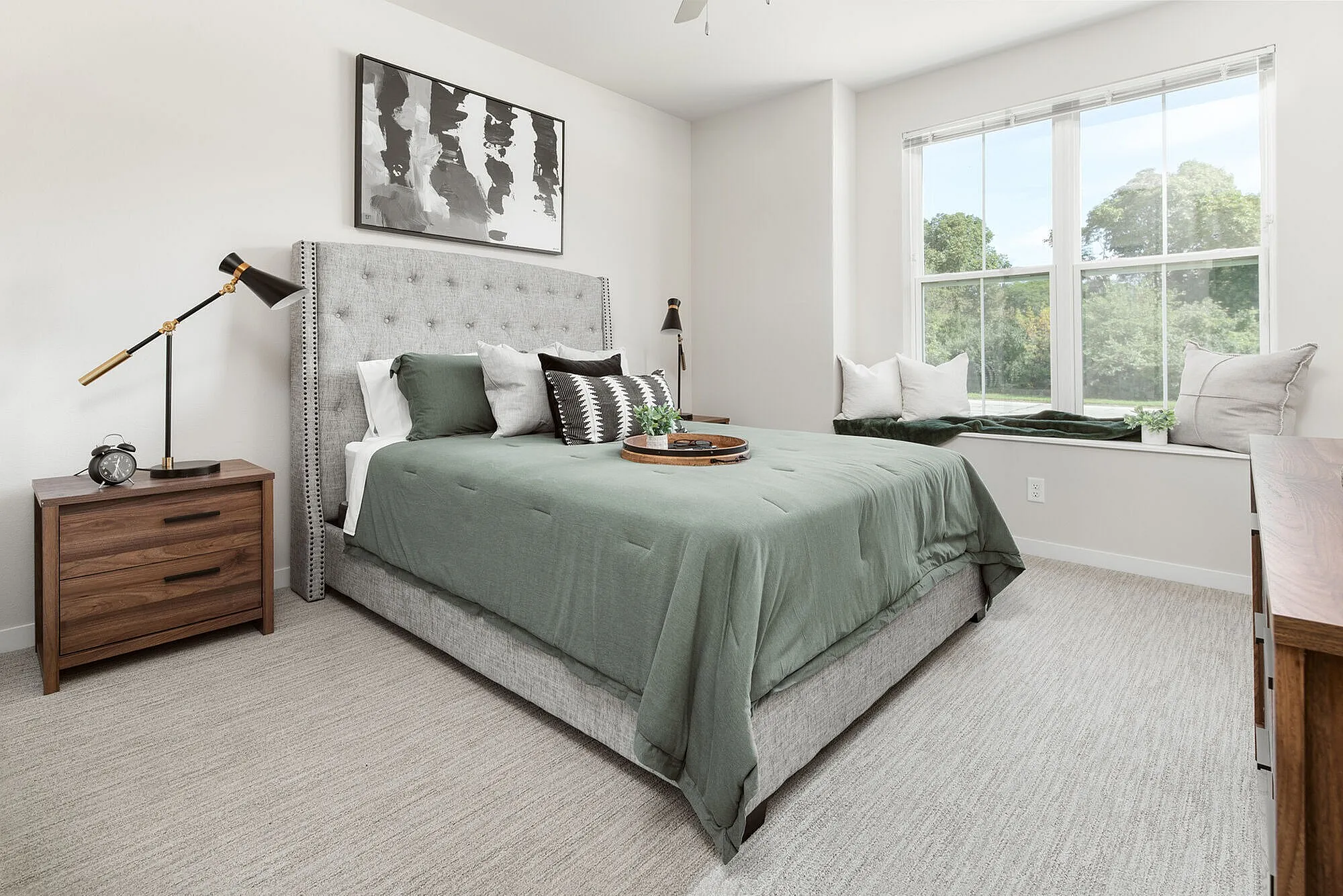 Bright and elegant bedroom with a plush gray bed, chic side tables, and large windows offering a serene view of greenery.