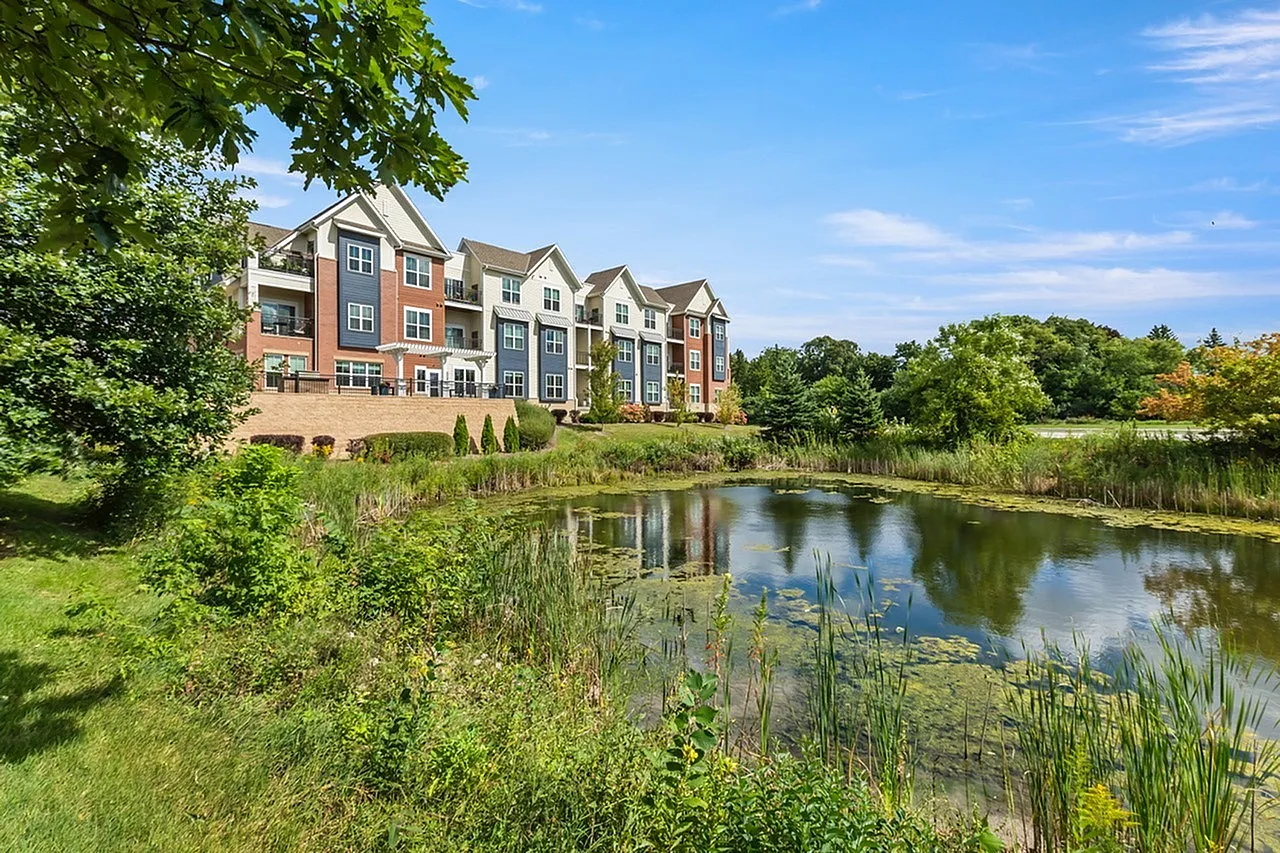 Charming apartment complex with colorful facades overlooking a serene pond and lush greenery under a clear blue sky.