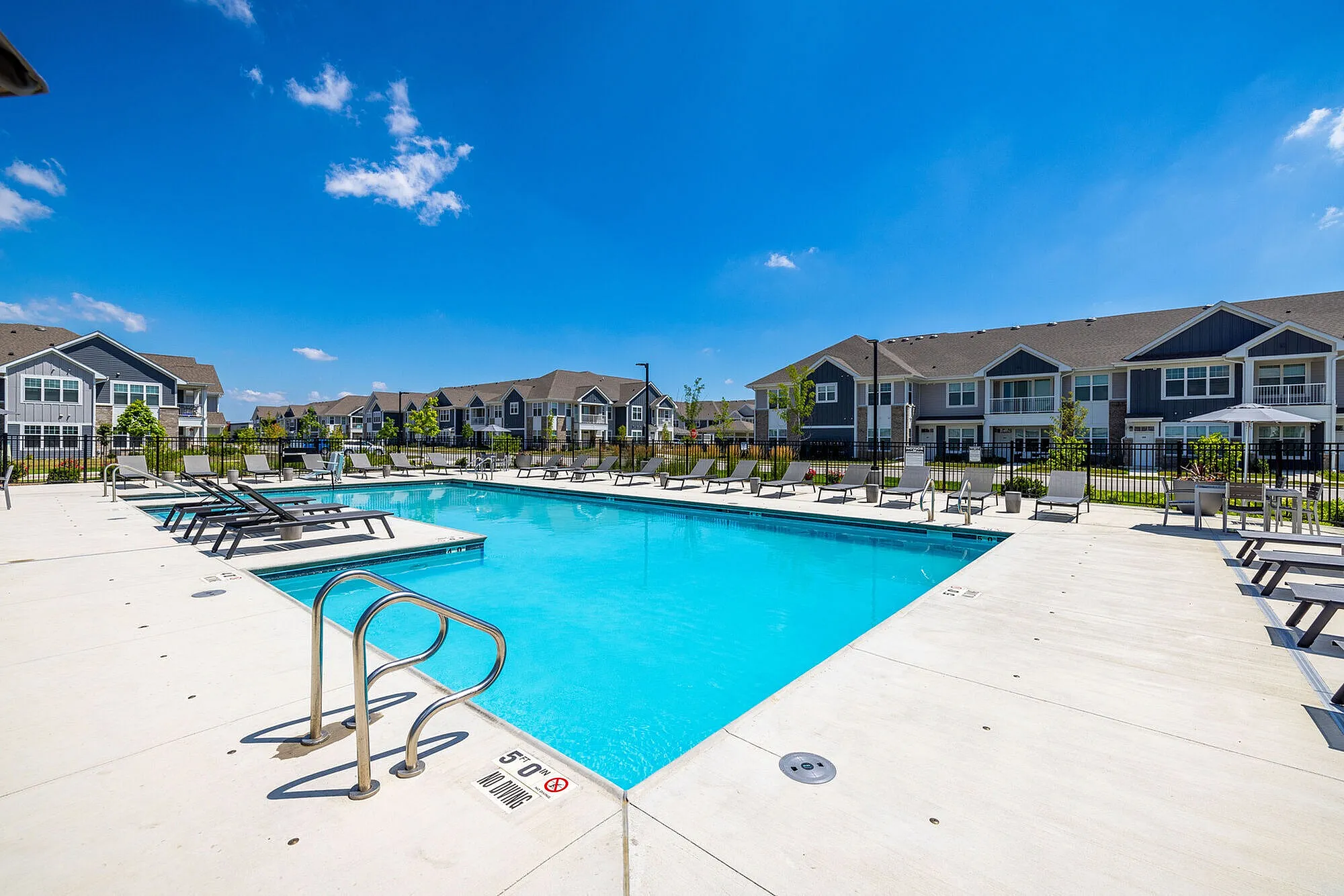 Inviting outdoor pool area with stylish lounge chairs and beautifully landscaped surroundings in a modern residential complex under a clear blue sky.