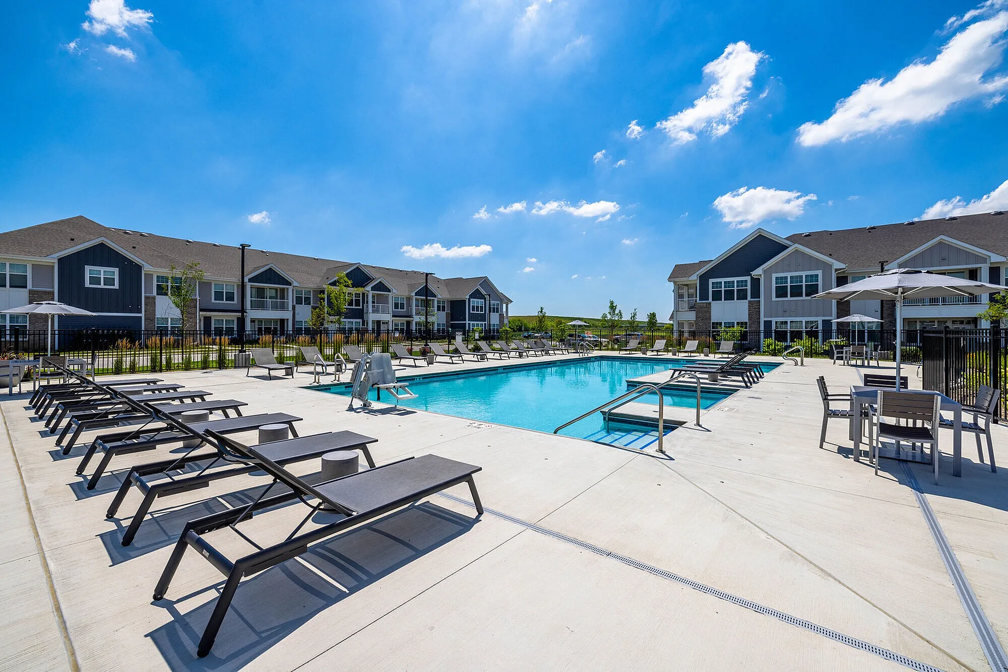 Luxurious apartment complex featuring a sparkling swimming pool surrounded by modern sun loungers, with charming two-story buildings in the background under a clear blue sky.