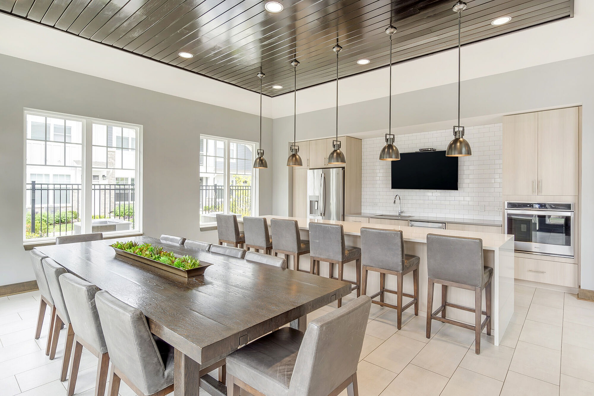 Modern communal dining area with elegant pendant lighting, large wooden table, and chic open-plan kitchen featuring stainless steel appliances and stylish countertops.