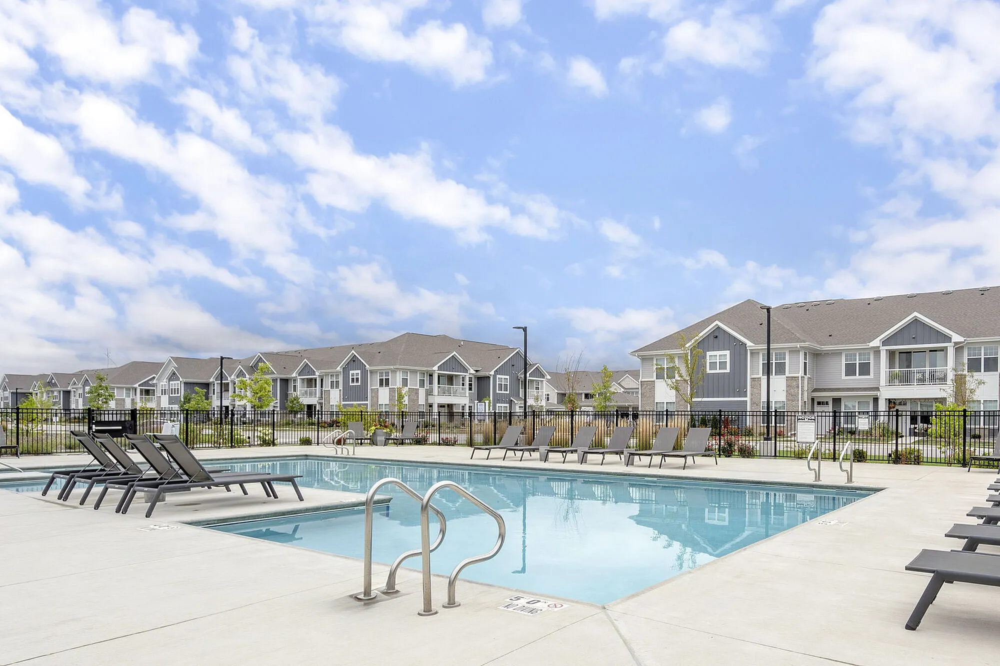 Modern apartment complex with inviting outdoor pool and sleek lounge chairs under a bright blue sky, offering a luxurious lifestyle.