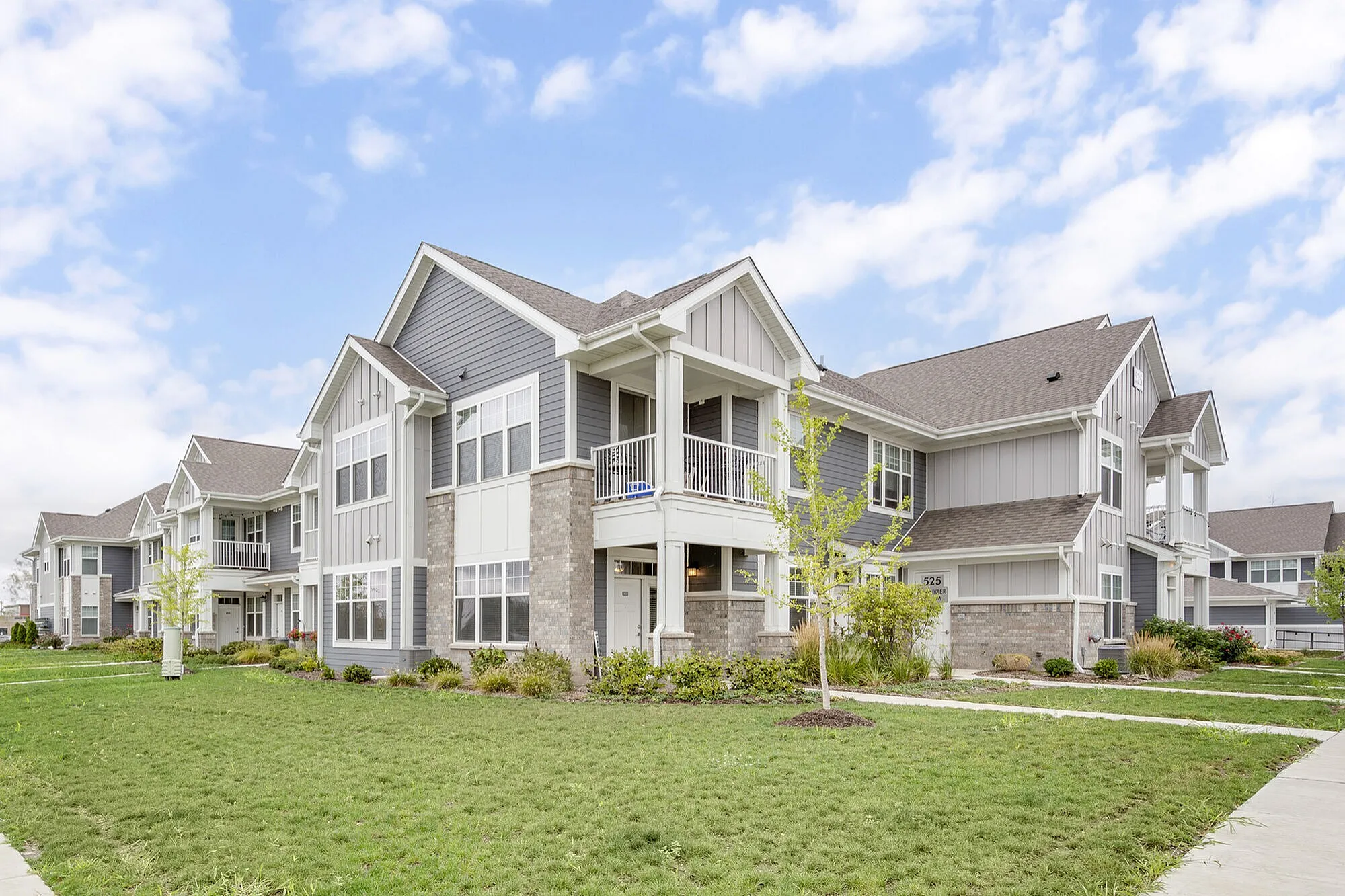Charming two-story apartment complex with modern architecture, featuring gray siding and stone accents, surrounded by lush greenery and a well-maintained lawn under a bright blue sky.