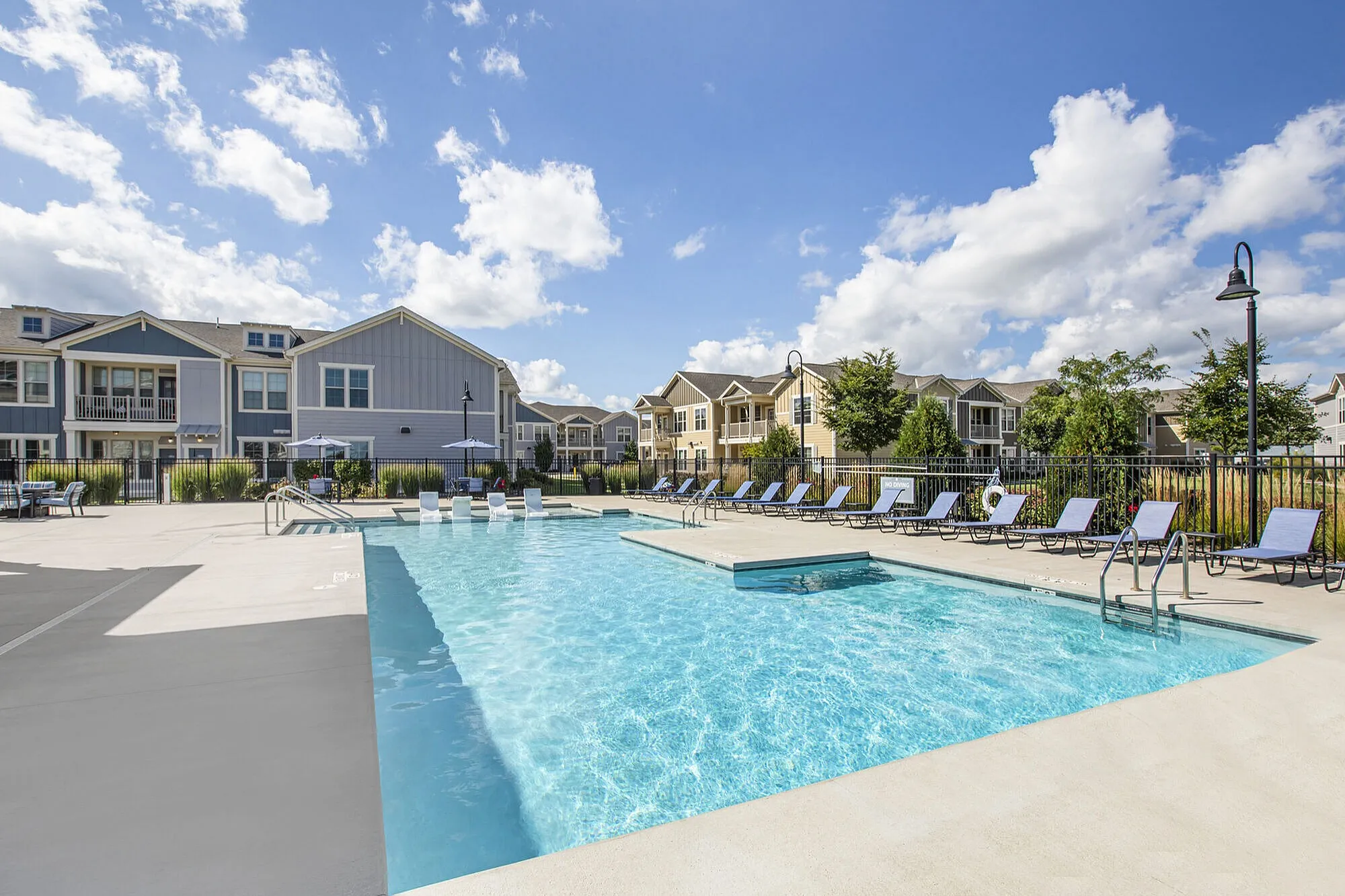 Modern apartment complex featuring a sparkling outdoor pool, surrounded by sun loungers and lush greenery under a bright blue sky.