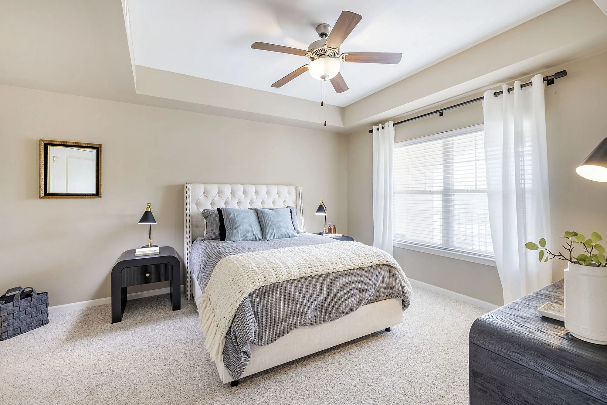 Bright and elegant bedroom featuring a plush upholstered bed, soft gray and white bedding, and a ceiling fan, illuminated by natural light from large windows with sheer curtains.