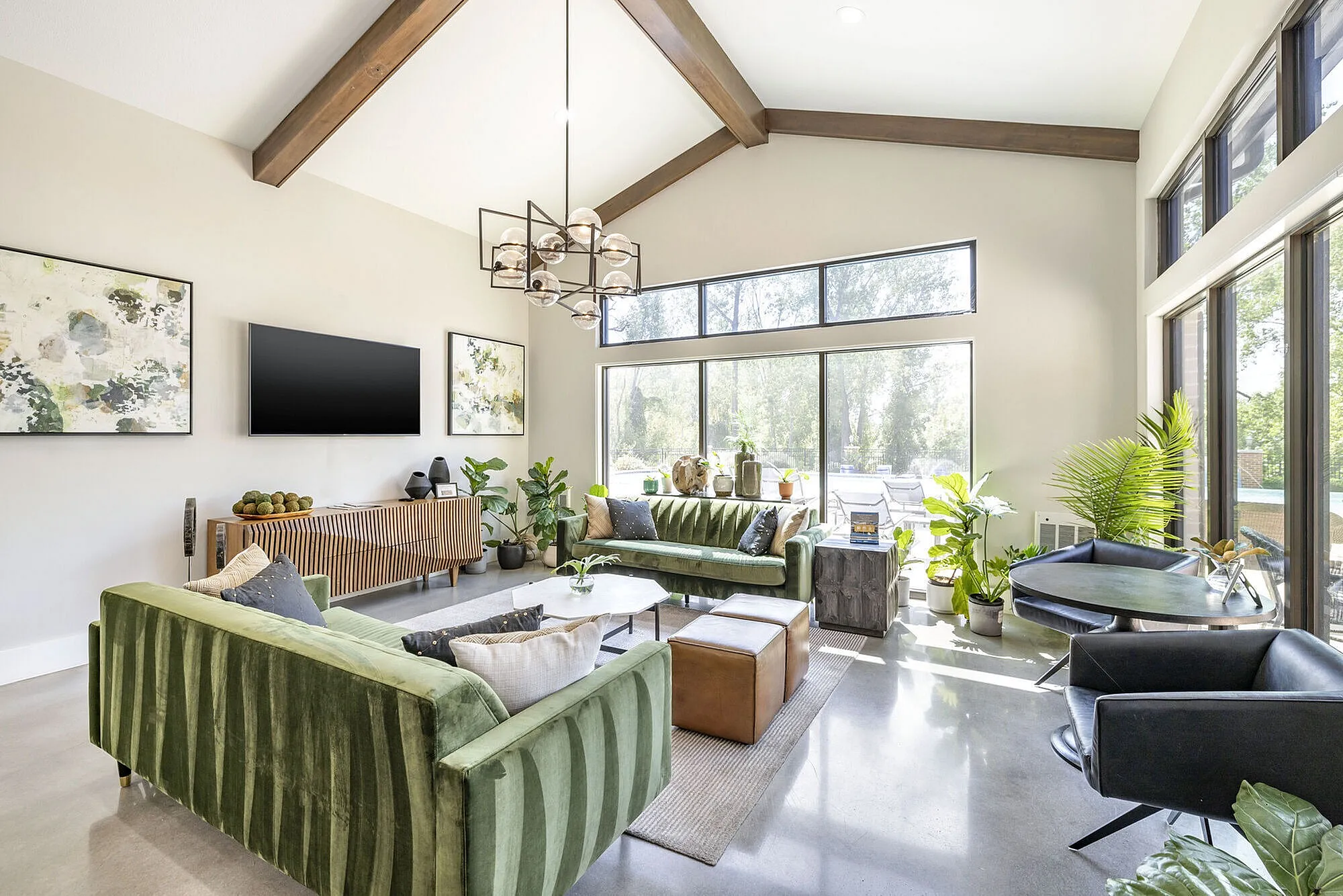 Bright and airy living room with modern decor, featuring green velvet sofas, high ceilings with wooden beams, and large windows overlooking lush greenery.