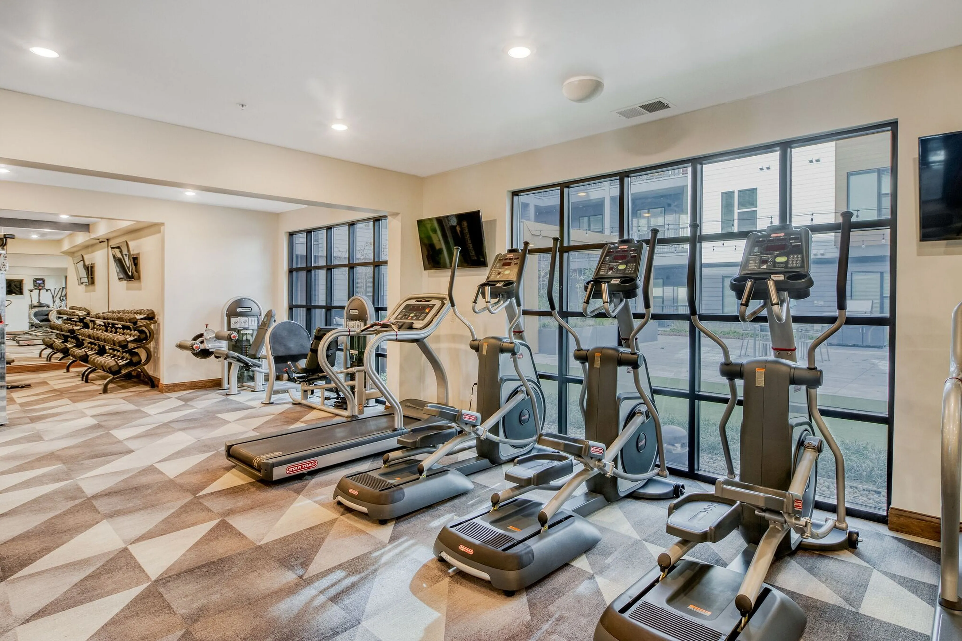 Modern fitness center featuring state-of-the-art exercise equipment, large windows with courtyard views, and a stylish geometric carpet design.