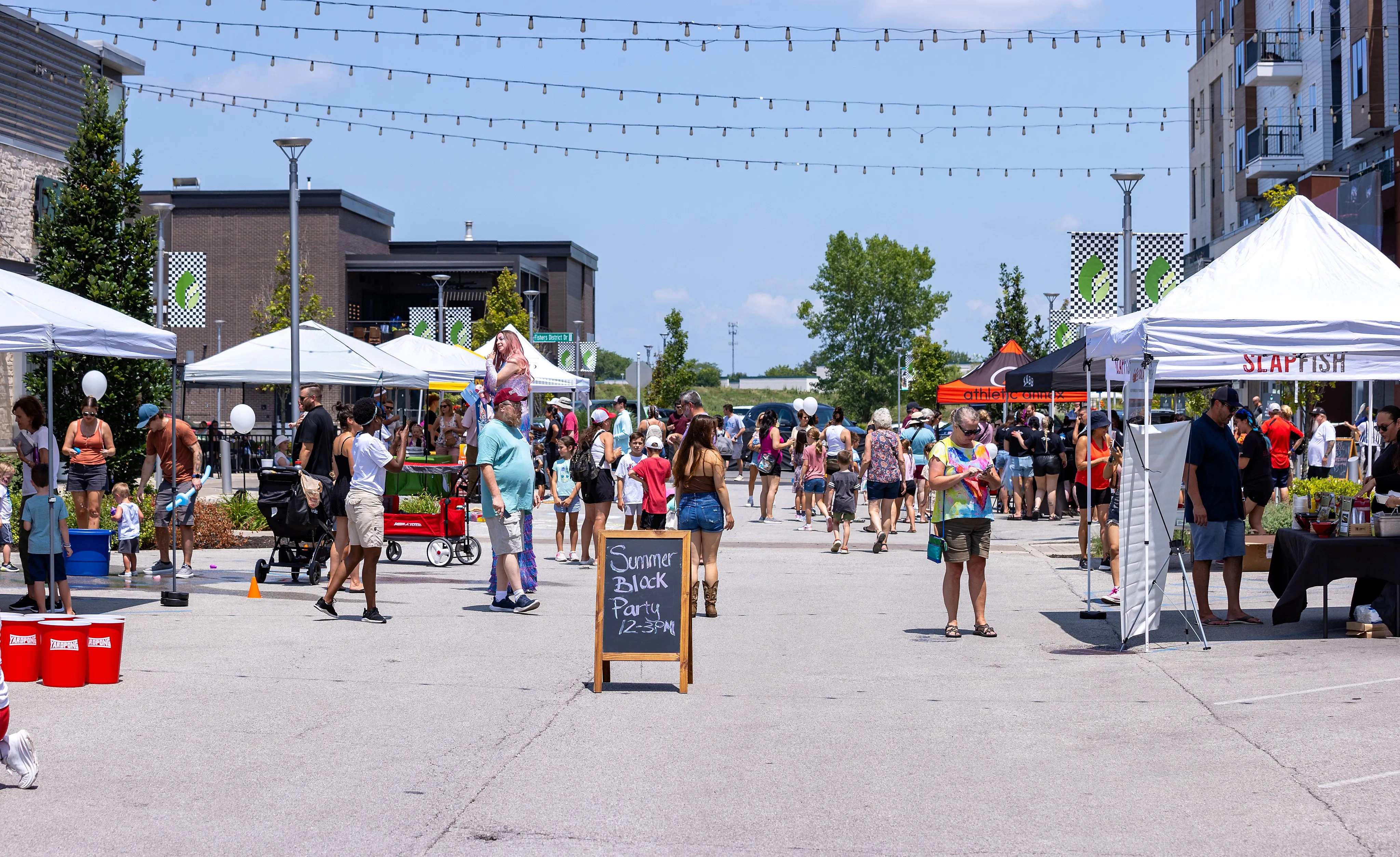 Vibrant summer block party with lively crowds, colorful tents, and festive decorations under a sunny sky.