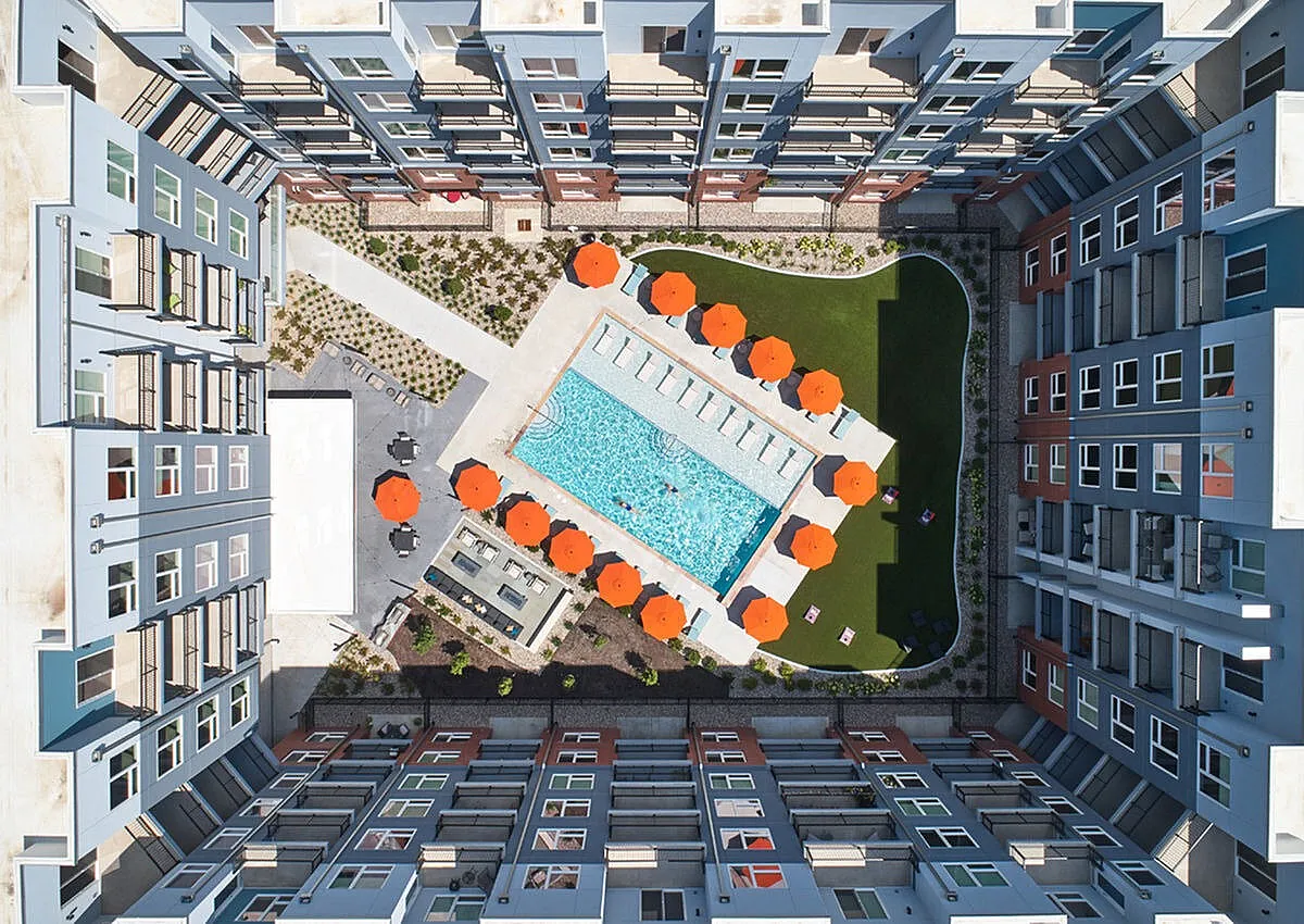 Stunning aerial view of modern apartment complex featuring a luxurious pool with vibrant orange umbrellas, surrounded by beautifully landscaped greenery and sleek architecture.
