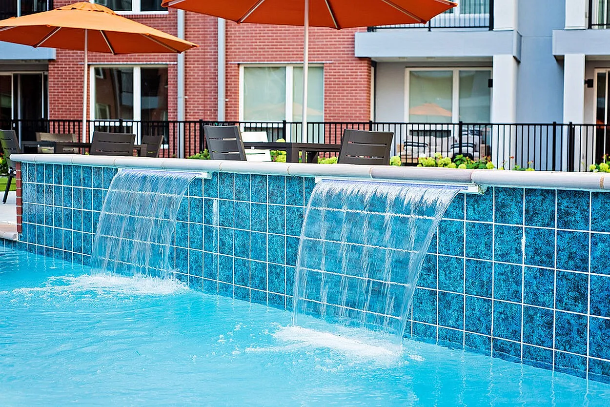 Luxurious apartment pool area featuring cascading waterfalls, vibrant blue tiles, and stylish outdoor seating with orange umbrellas.