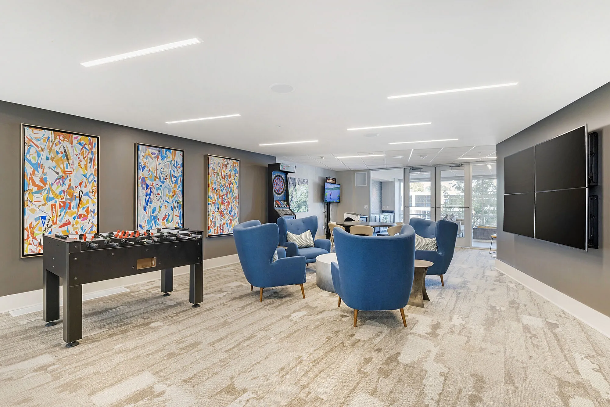 Modern recreation room with vibrant artwork, a foosball table, and plush blue seating, featuring large windows for natural light.