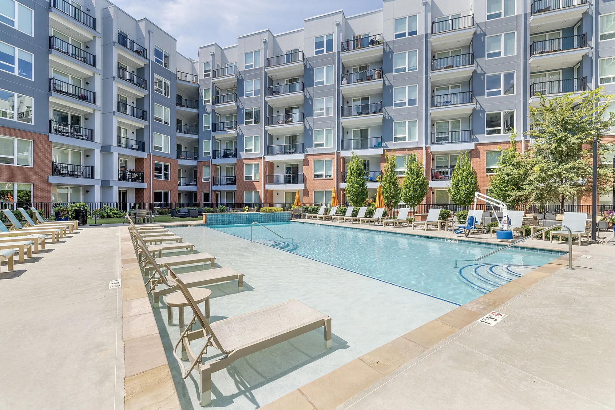 Luxurious modern apartment complex with a sparkling blue swimming pool, surrounded by elegant sun loungers and vibrant greenery.