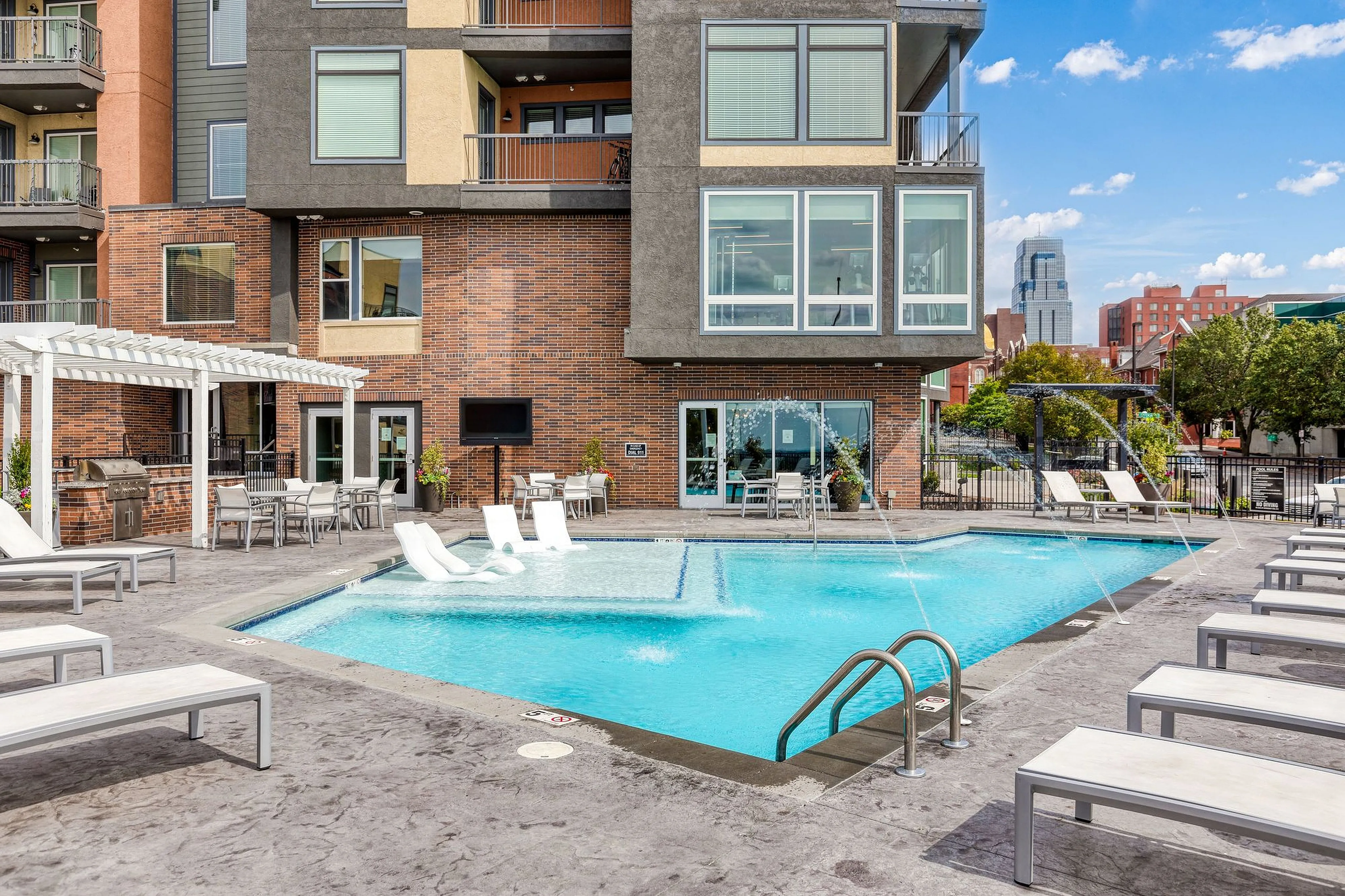 Modern apartment complex featuring a stylish pool with water features, surrounded by lounge chairs and outdoor seating under a pergola.