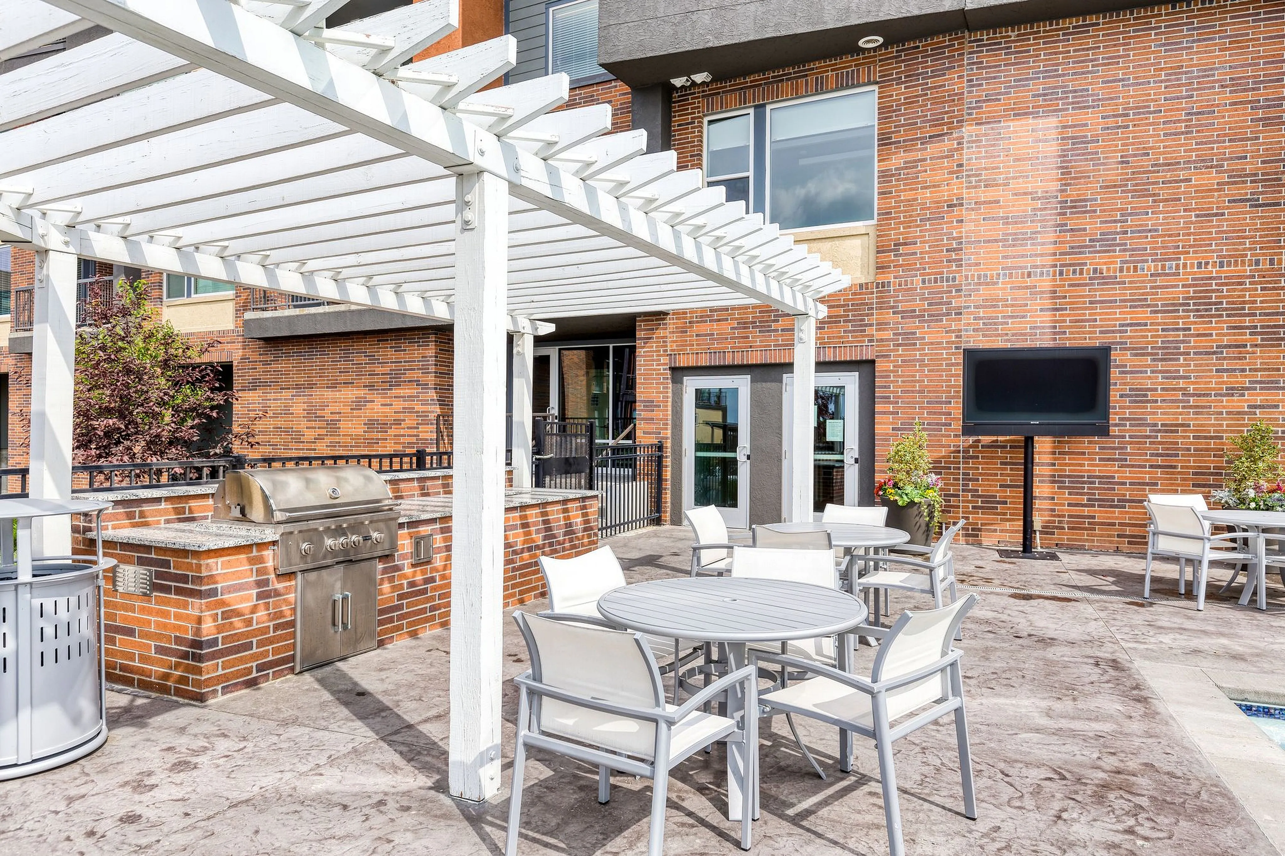 Modern outdoor patio featuring a barbecue grill, elegant seating area with white tables and chairs, and lush greenery against a stylish brick building backdrop.