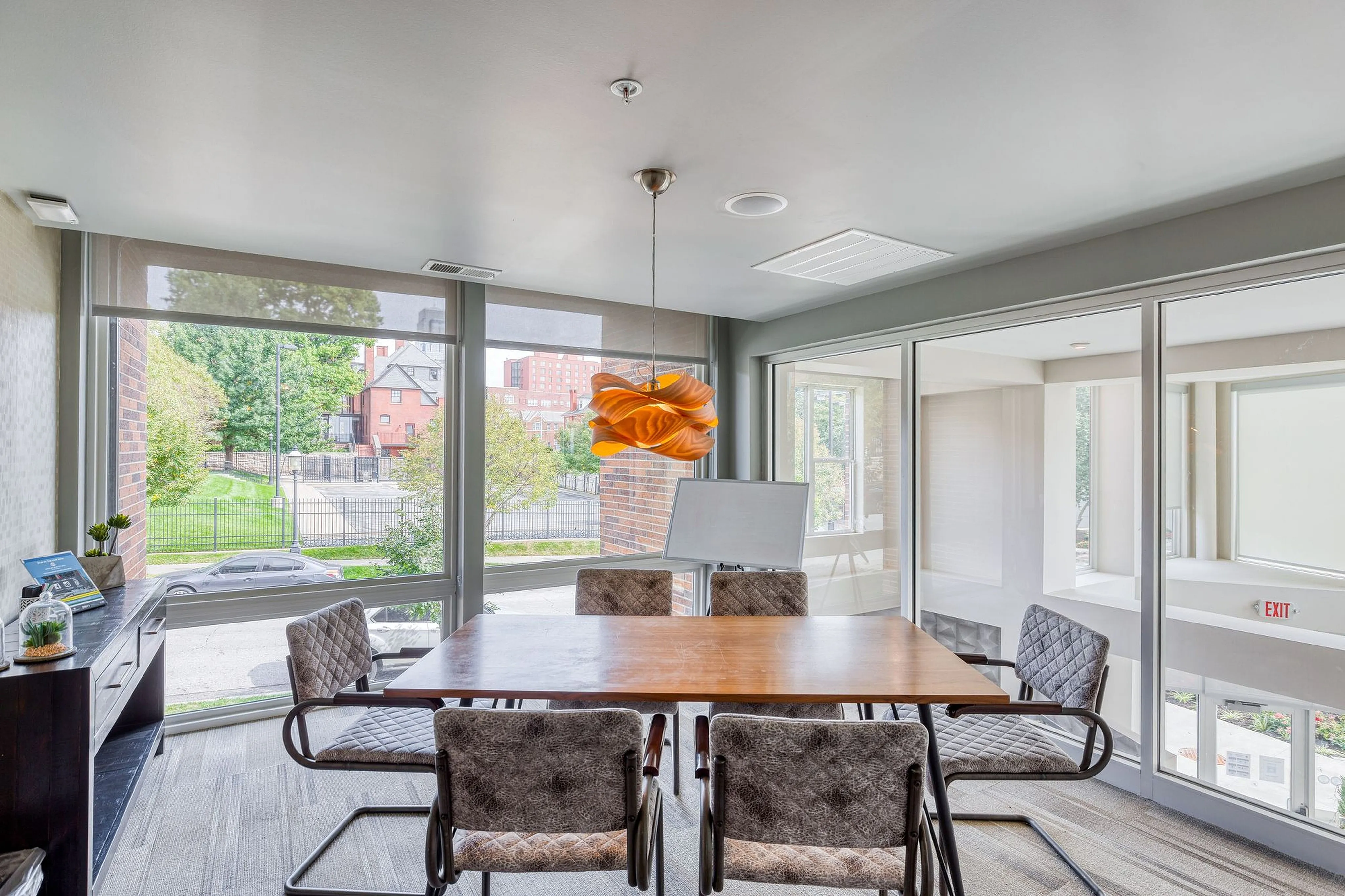 Modern conference room with a large wooden table, stylish chairs, and a unique orange pendant light, offering a vibrant and spacious atmosphere with large windows and a view of the surrounding neighborhood.