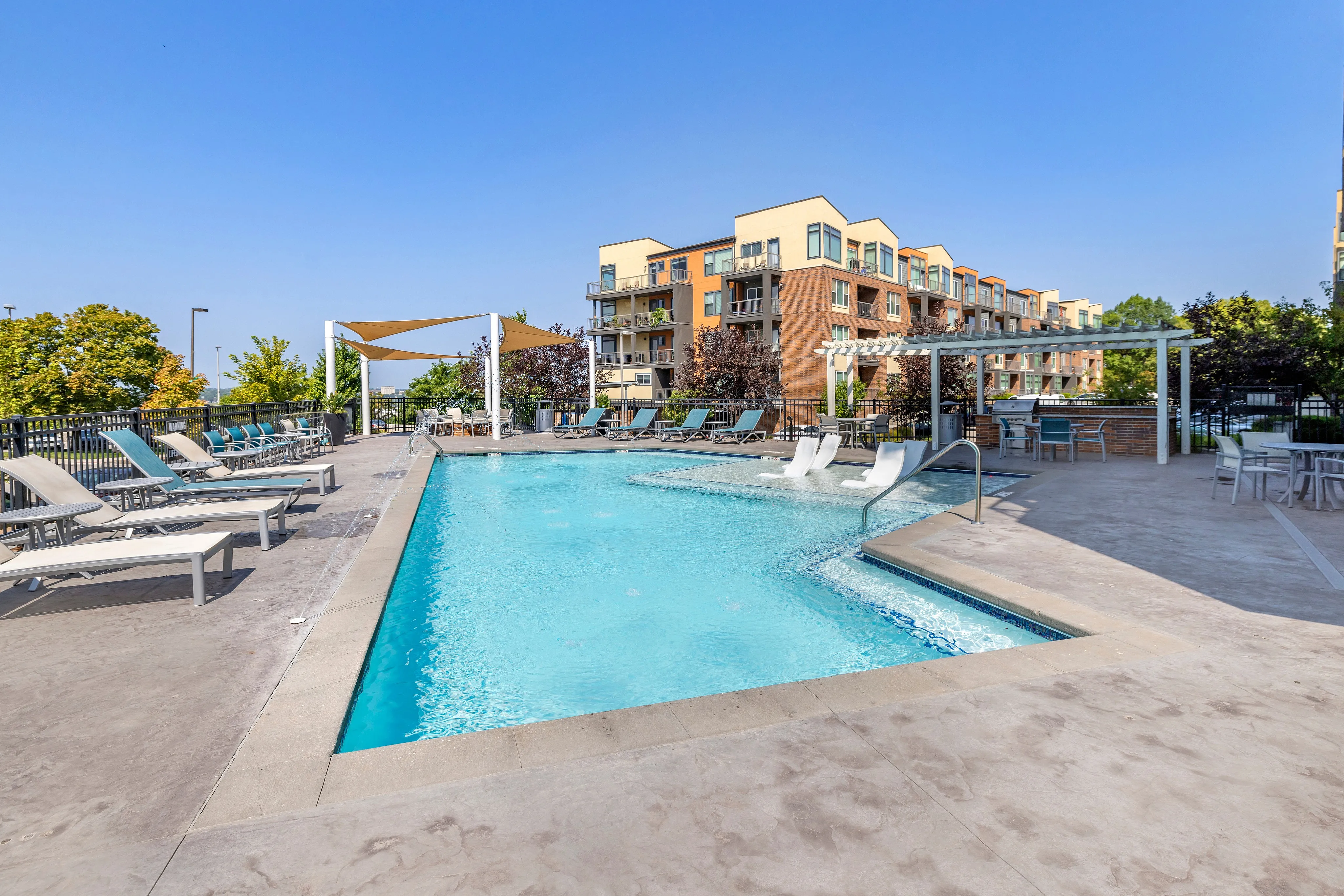 Inviting outdoor pool area with luxurious lounge chairs and shaded seating, perfect for relaxation and social gatherings at a modern apartment complex.