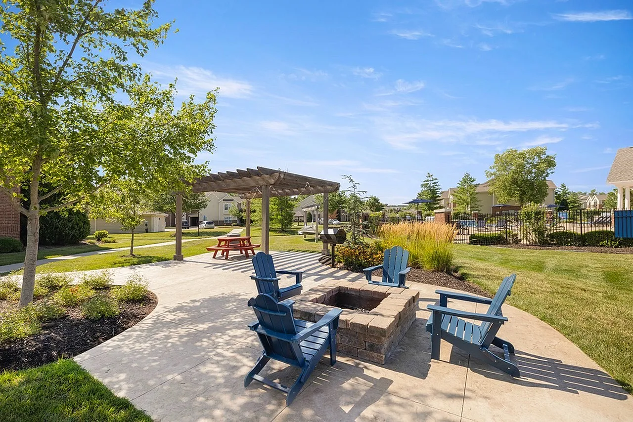 Charming outdoor space with a cozy fire pit, blue Adirondack chairs, a picnic table under a pergola, and lush landscaping, ideal for relaxation and gatherings.