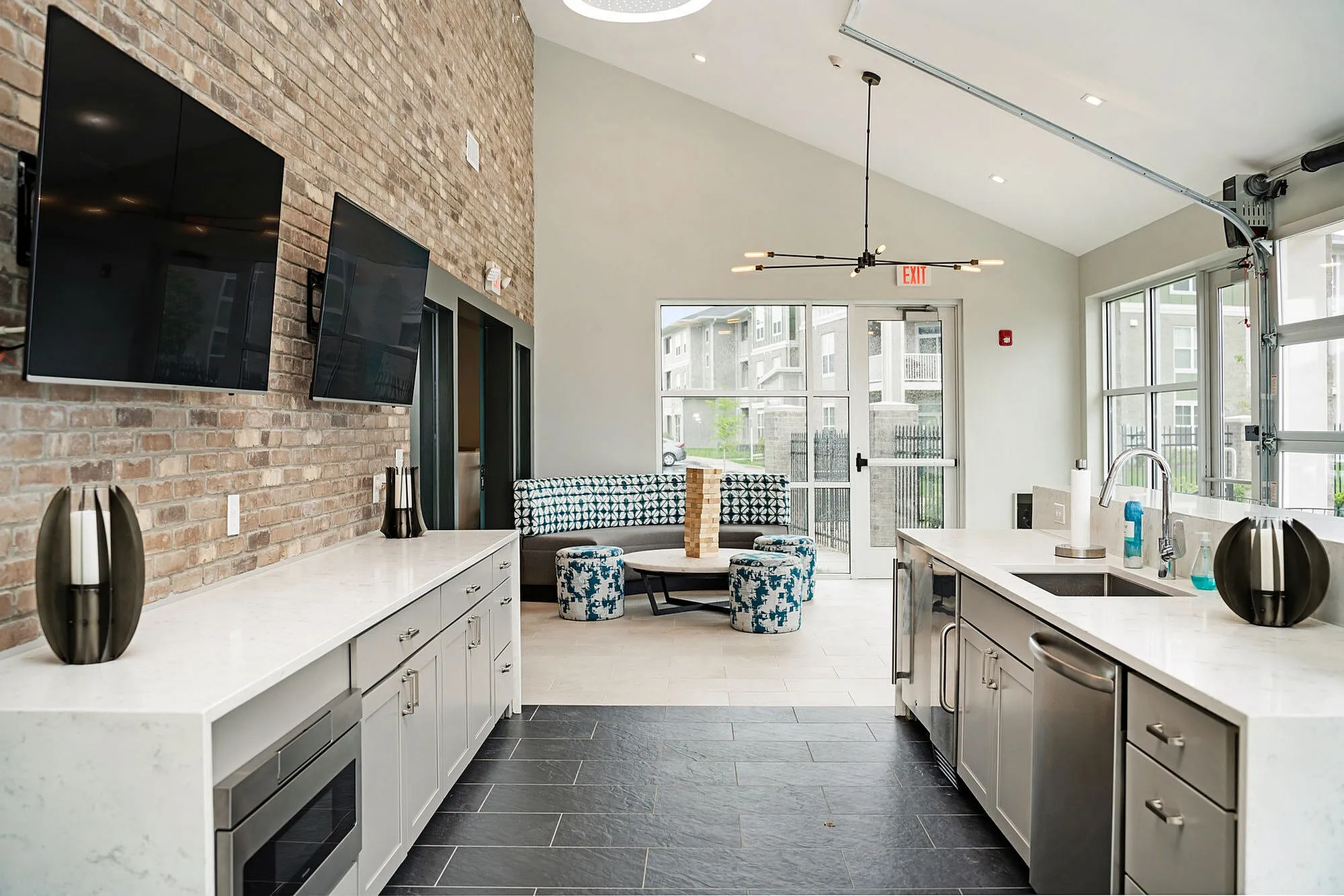 Modern communal area with sleek kitchen, dual TVs mounted on a stylish brick wall, and a cozy seating area with natural light streaming through large windows.