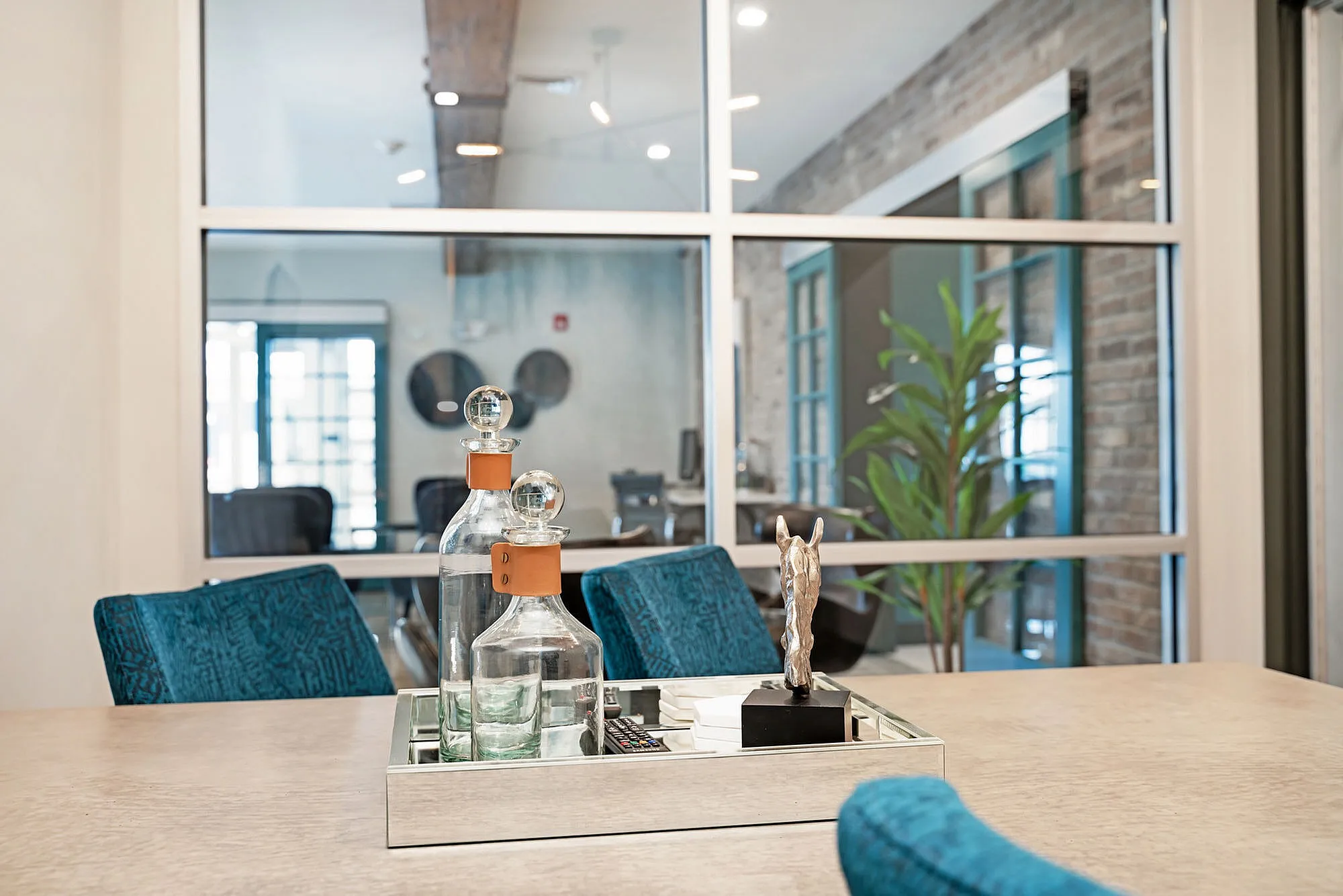 Elegant modern room featuring a stylish decor tray with glass bottles and horse statuette, complemented by teal chairs and abundant natural light from spacious windows.