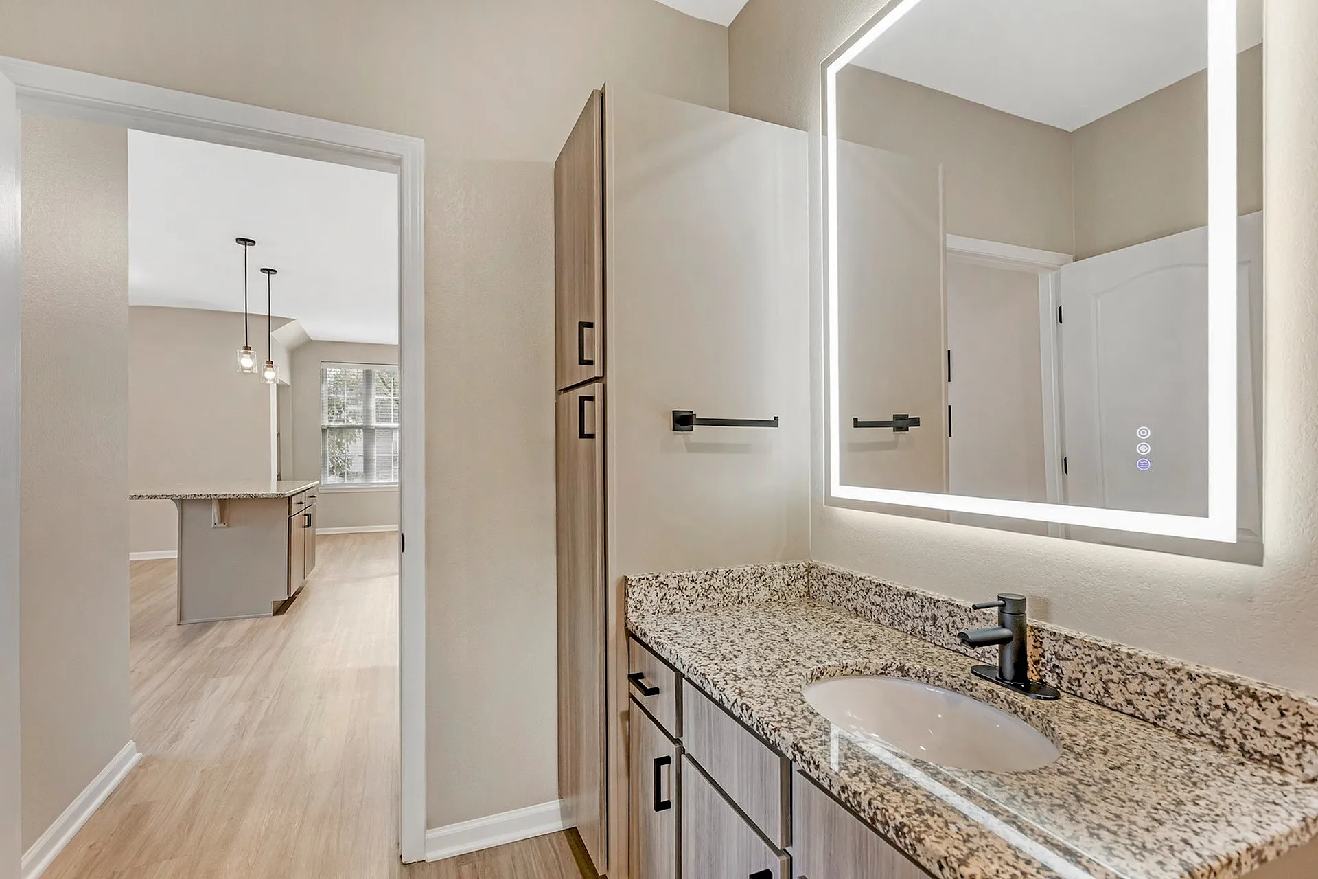 Elegant bathroom featuring a granite countertop and illuminated mirror, opening into a stylish open-plan kitchen with pendant lighting and modern cabinetry.