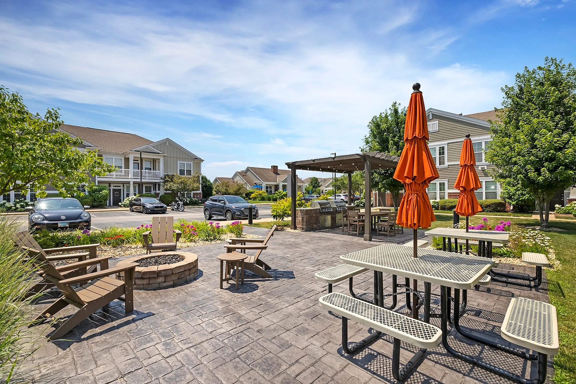 Inviting outdoor patio area with seating, vibrant orange umbrellas, and a cozy fire pit surrounded by lush greenery and modern apartment buildings.