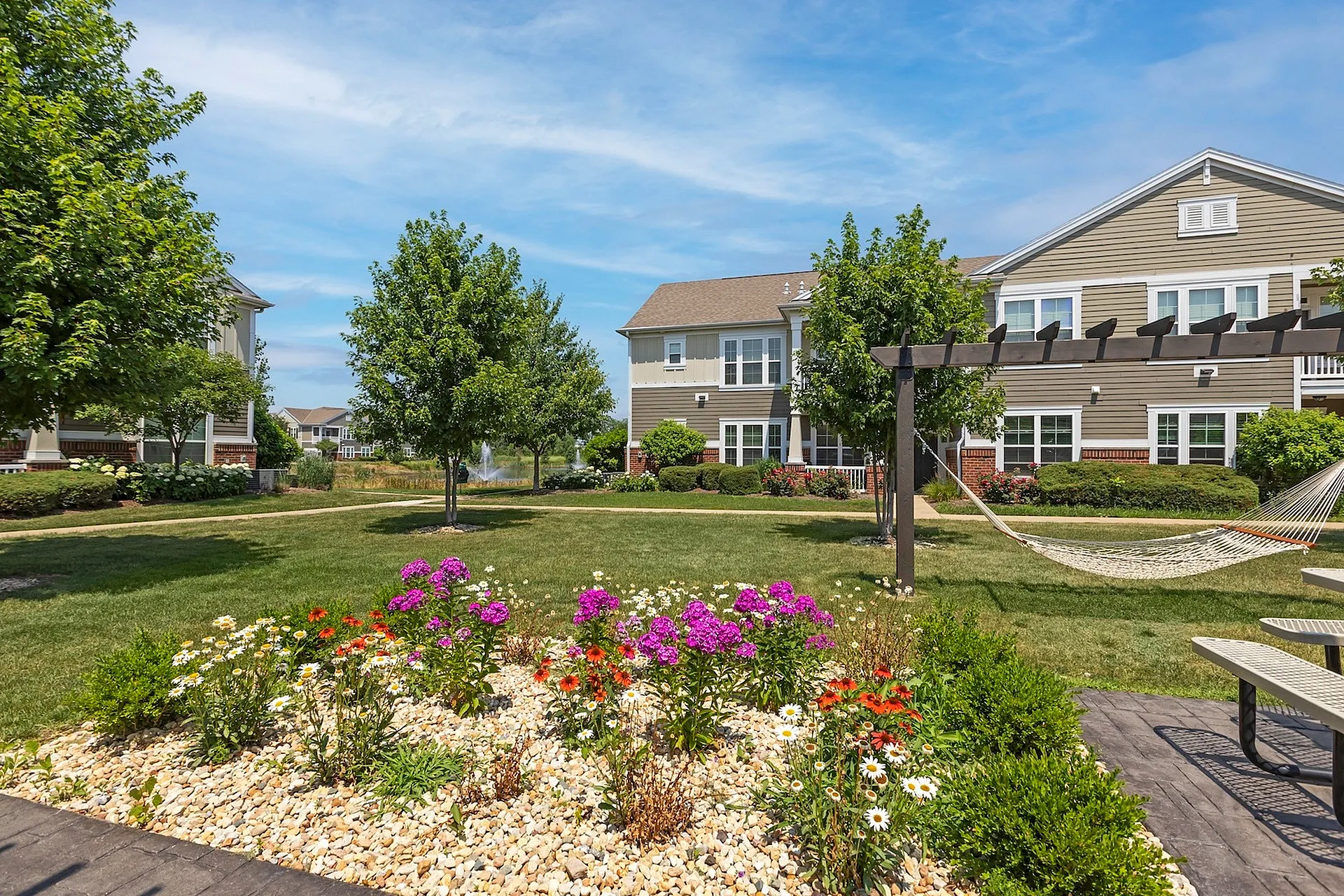 Charming garden view with vibrant flowers and mature trees set against a modern apartment complex, featuring a peaceful hammock and inviting picnic area.