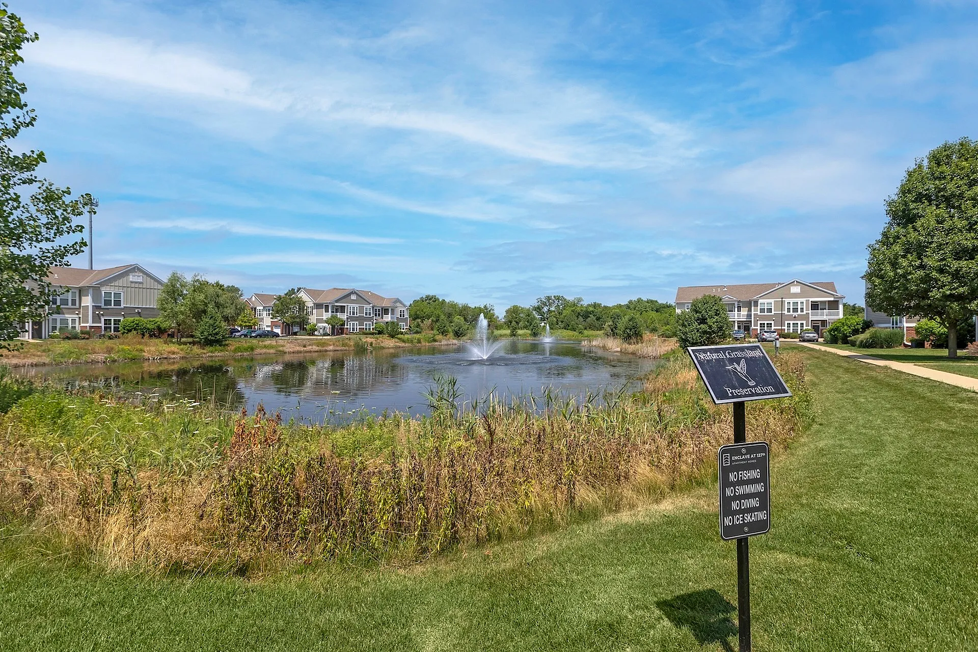 Charming residential community featuring serene ponds with elegant fountains, lush greenery, and spacious, modern apartments under a clear blue sky.